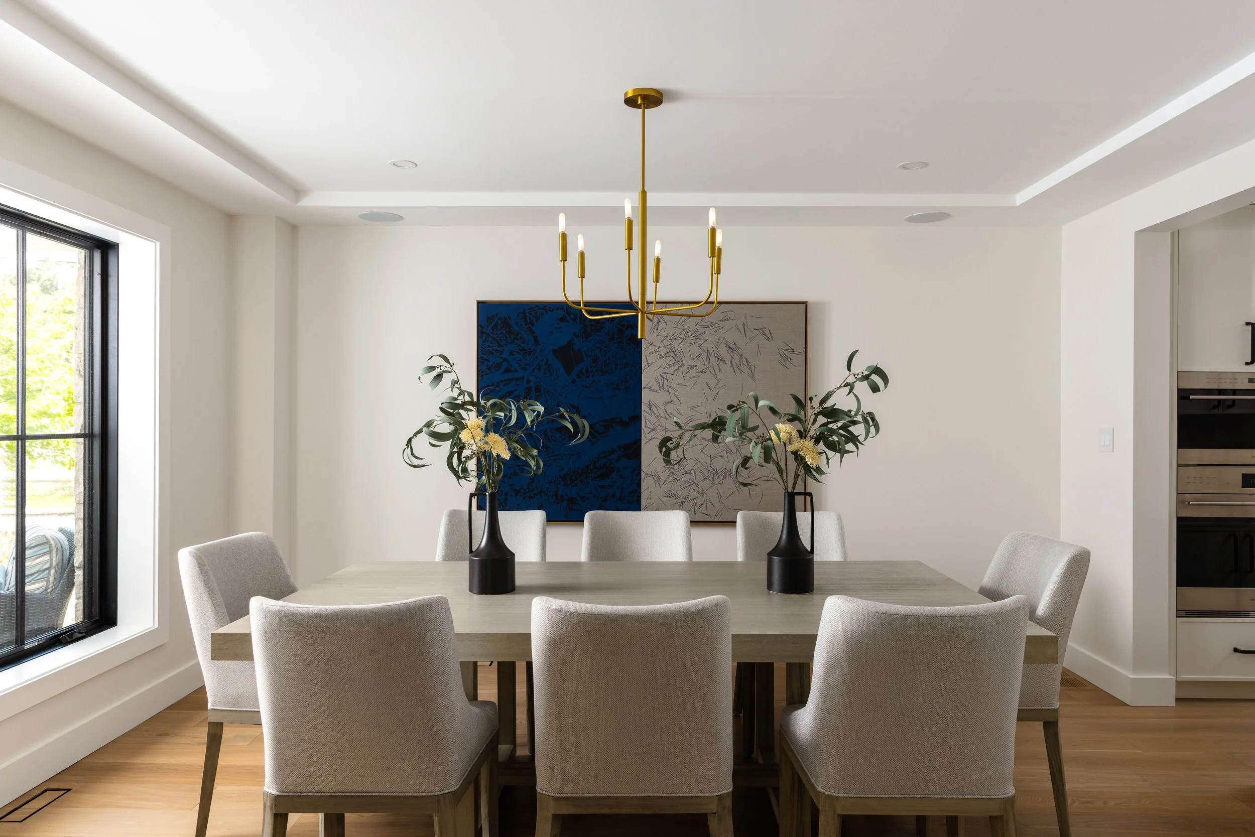 A modern dining room table with 8 white chairs, a modern candelabra-style light fixture overhead and a piece of grey and blue modern art on the wall