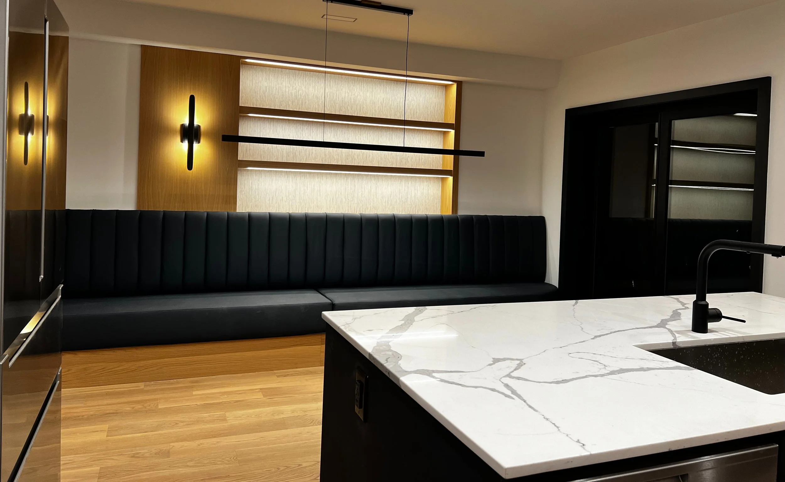 Long black upholstered banquette runs along the far wall, with custom shelving above and a wall sconce on the left side above the banquette. The corner of a white marble-topped kitchen island is in the foreground, with bare oak flooring in between.