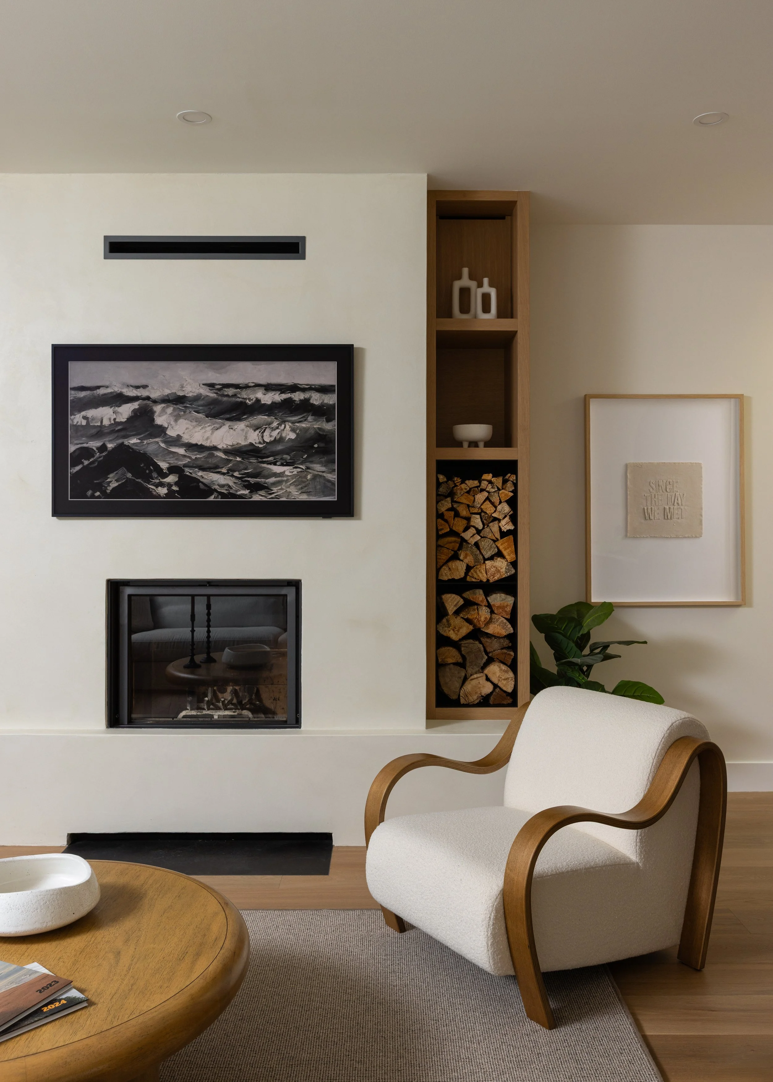 Close shot of wood-burning fireplace with flatscreen television hanging above, custom millwork shelving for firewood storage and a modern chair in white with wood accents