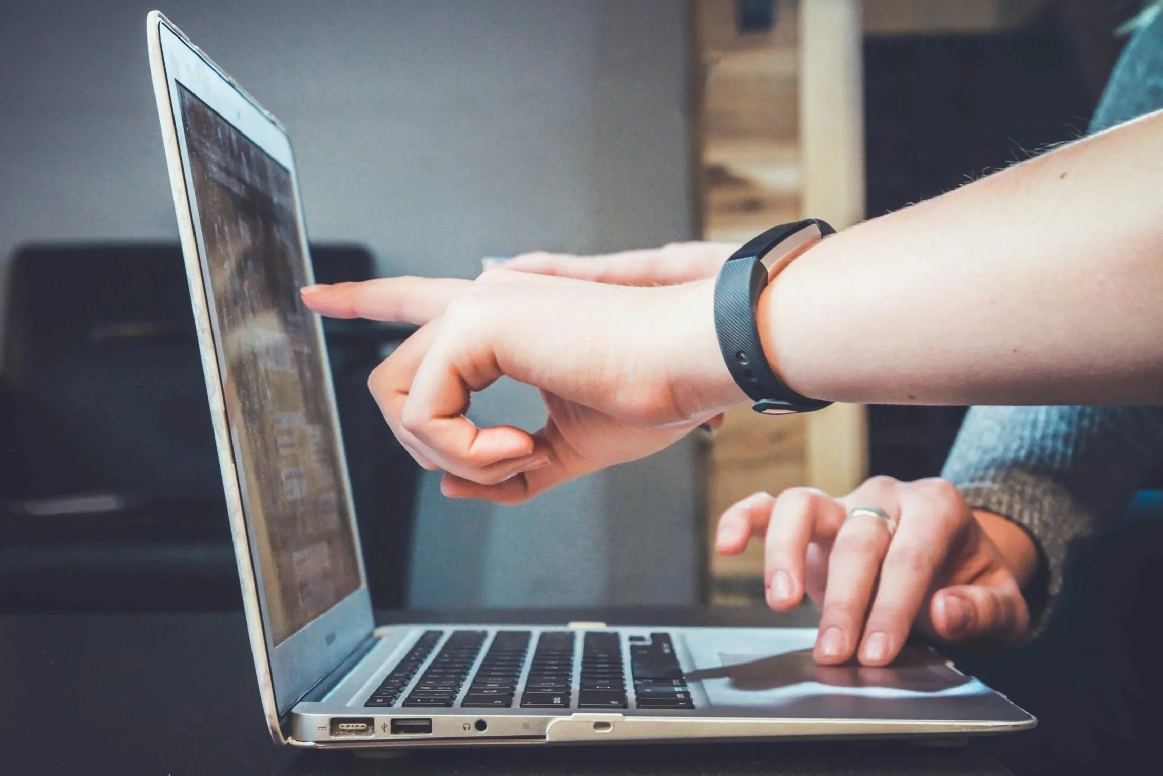 Person pointing at a laptop screen with another person watching, wearing a black wristband.