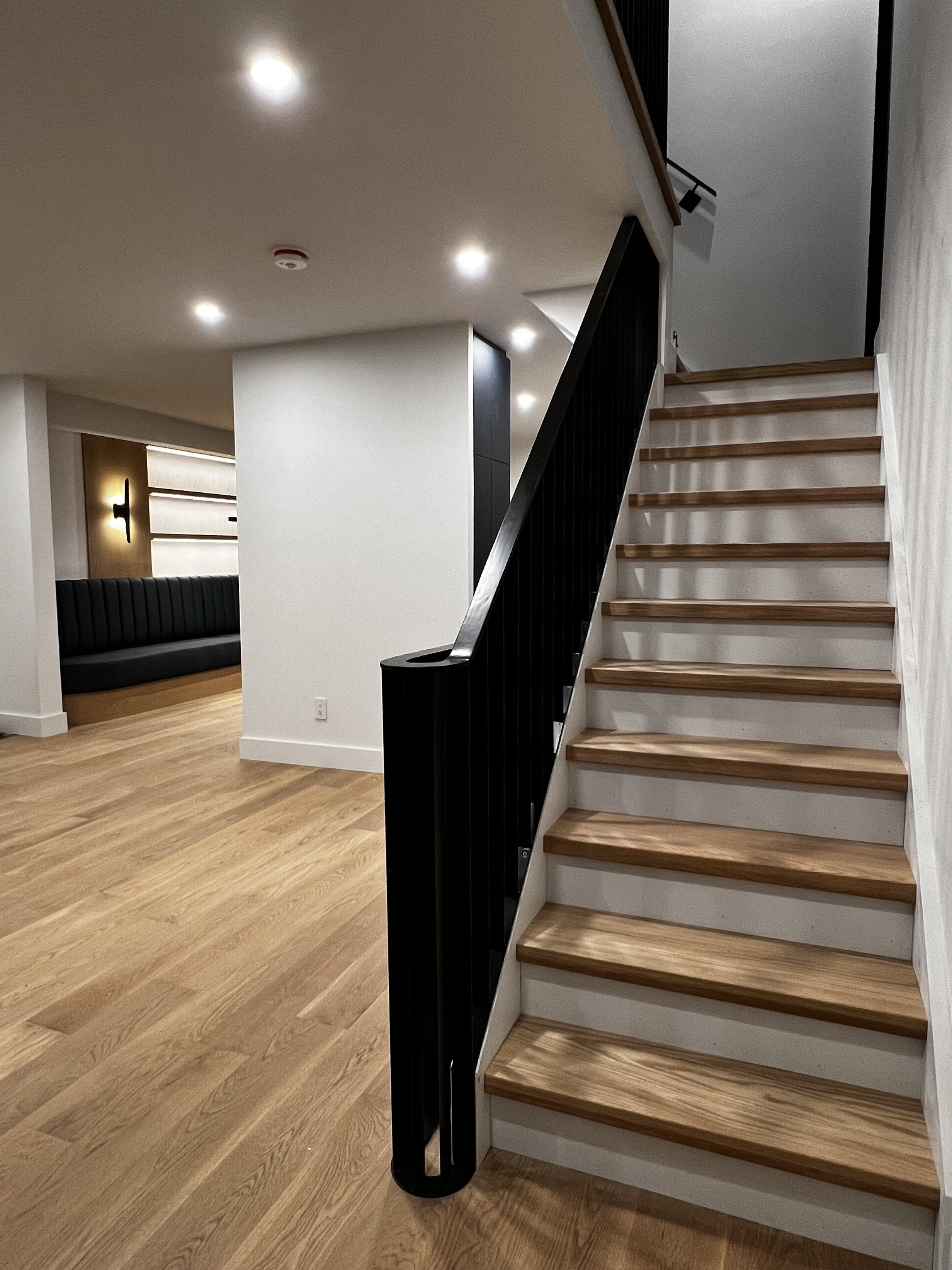 Stairwell as seen from front entryway. Oak flooring, black metal railing, white walls and oak stair treads are in the foreground and a black, upholstered banquette can be seen through the doorway to the kitchen