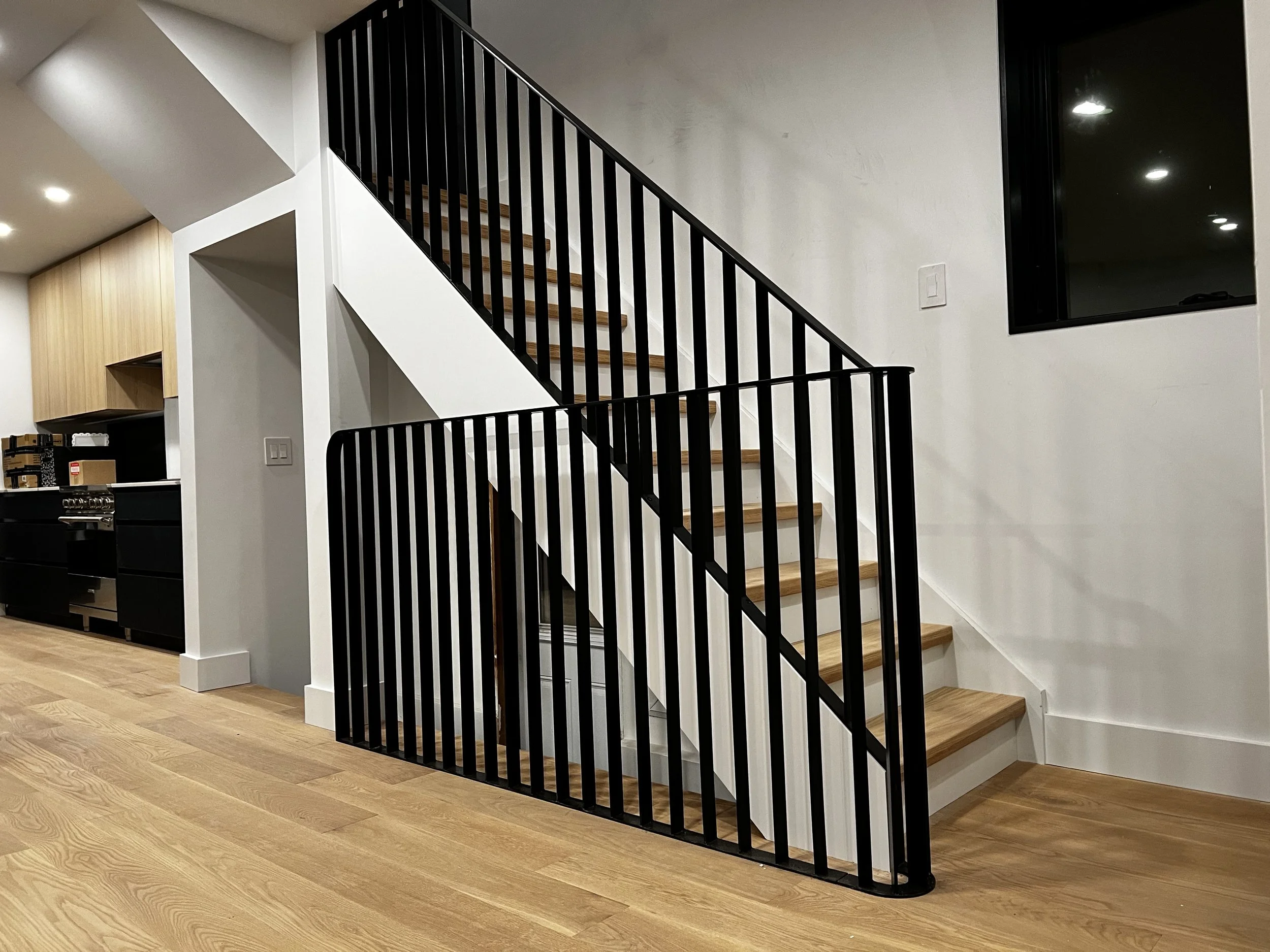 Sleek black metal railing and stair guard wrapping around white stairs with oak treads. Crisp white walls and pale oak hardwood flooring frame the stairwell