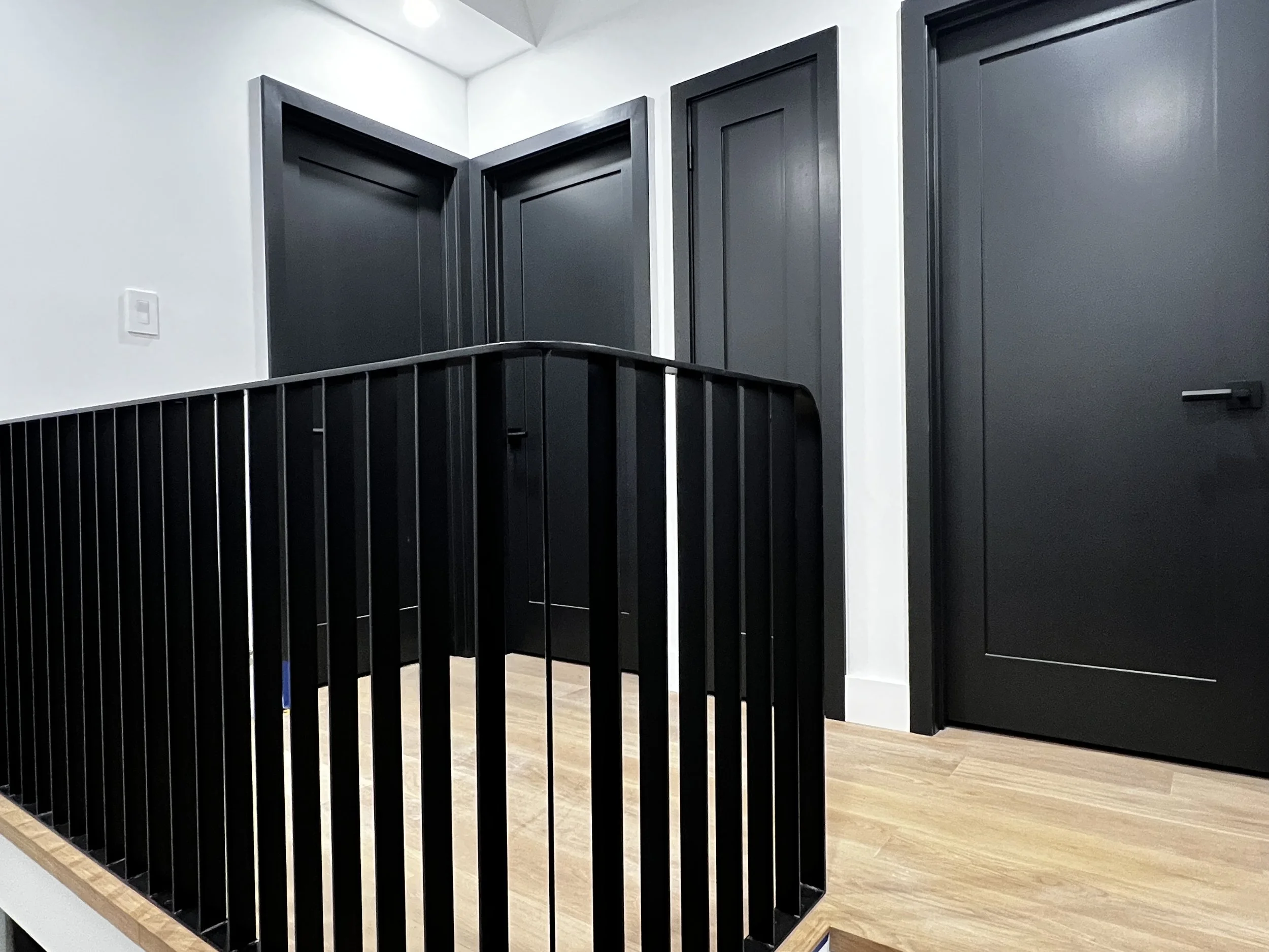 Upstairs hallway featuring custom black metal railing and stair guard, sleek black bedroom doors, oak flooring and white walls