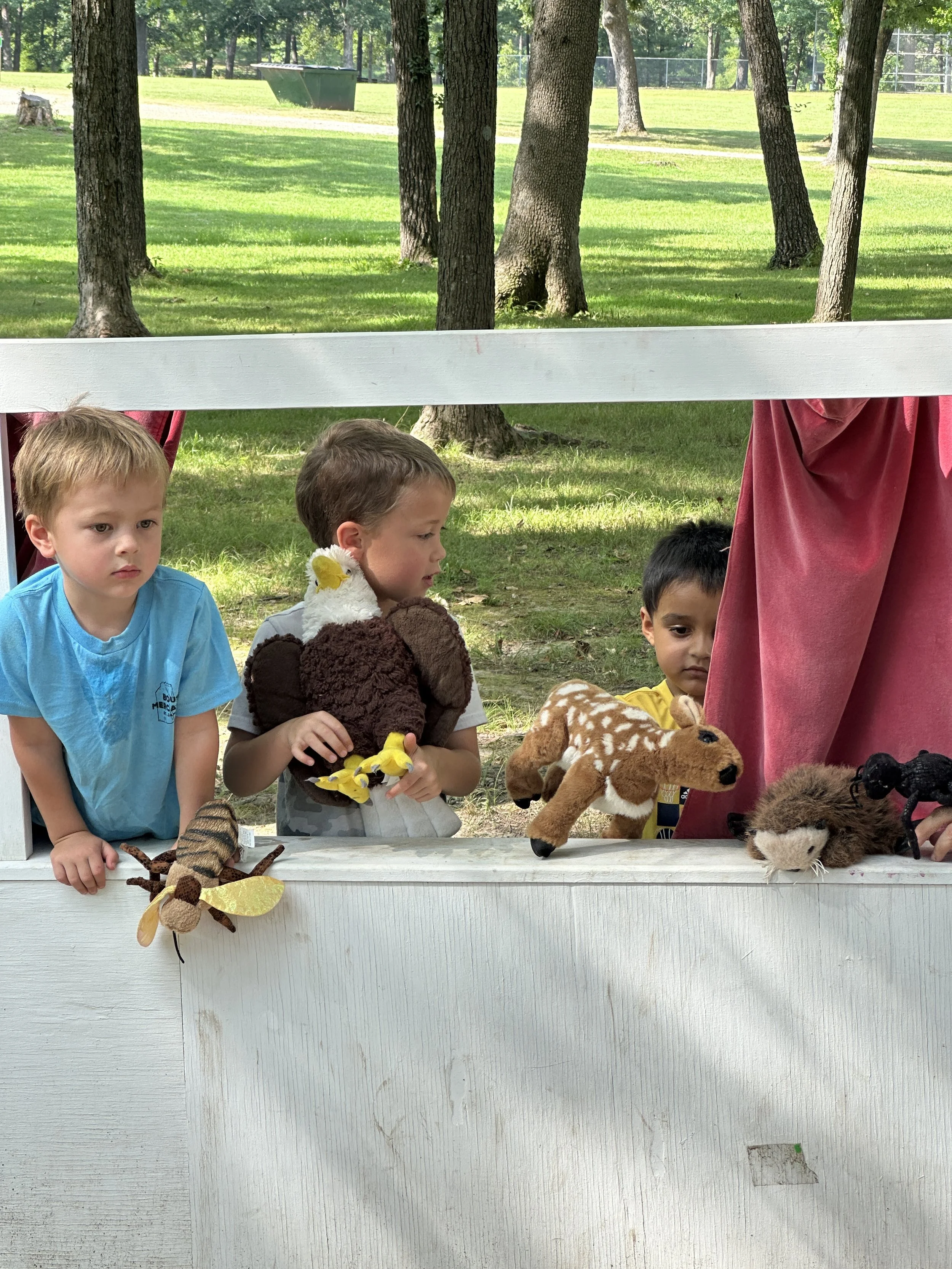 Three children looking through a white puppet theater. They are holding various plush animal puppets, including a bee, a goose, a horse, and a mouse. The background features a grassy park with trees.