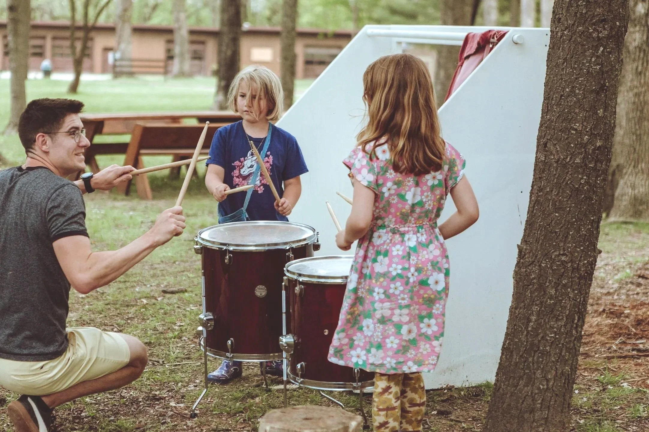 A man and two young girls playing drums outdoors in a park surrounded by trees.