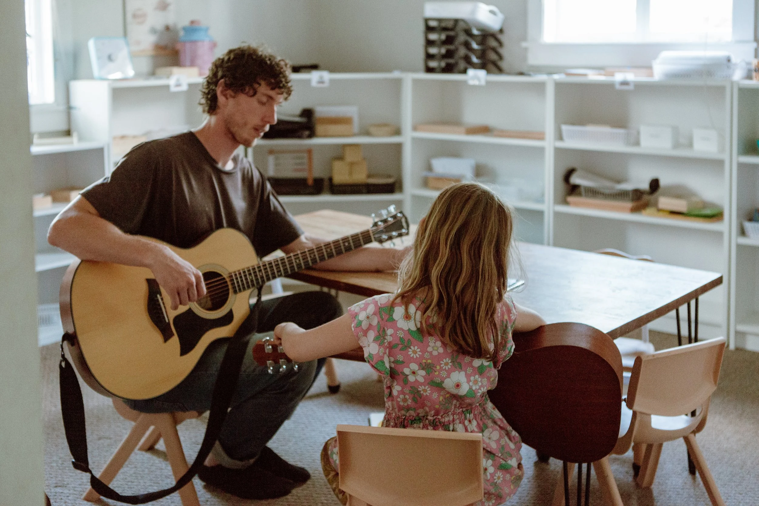 A young man playing an acoustic guitar for a young girl sitting at a table in a bright room with shelves and books.