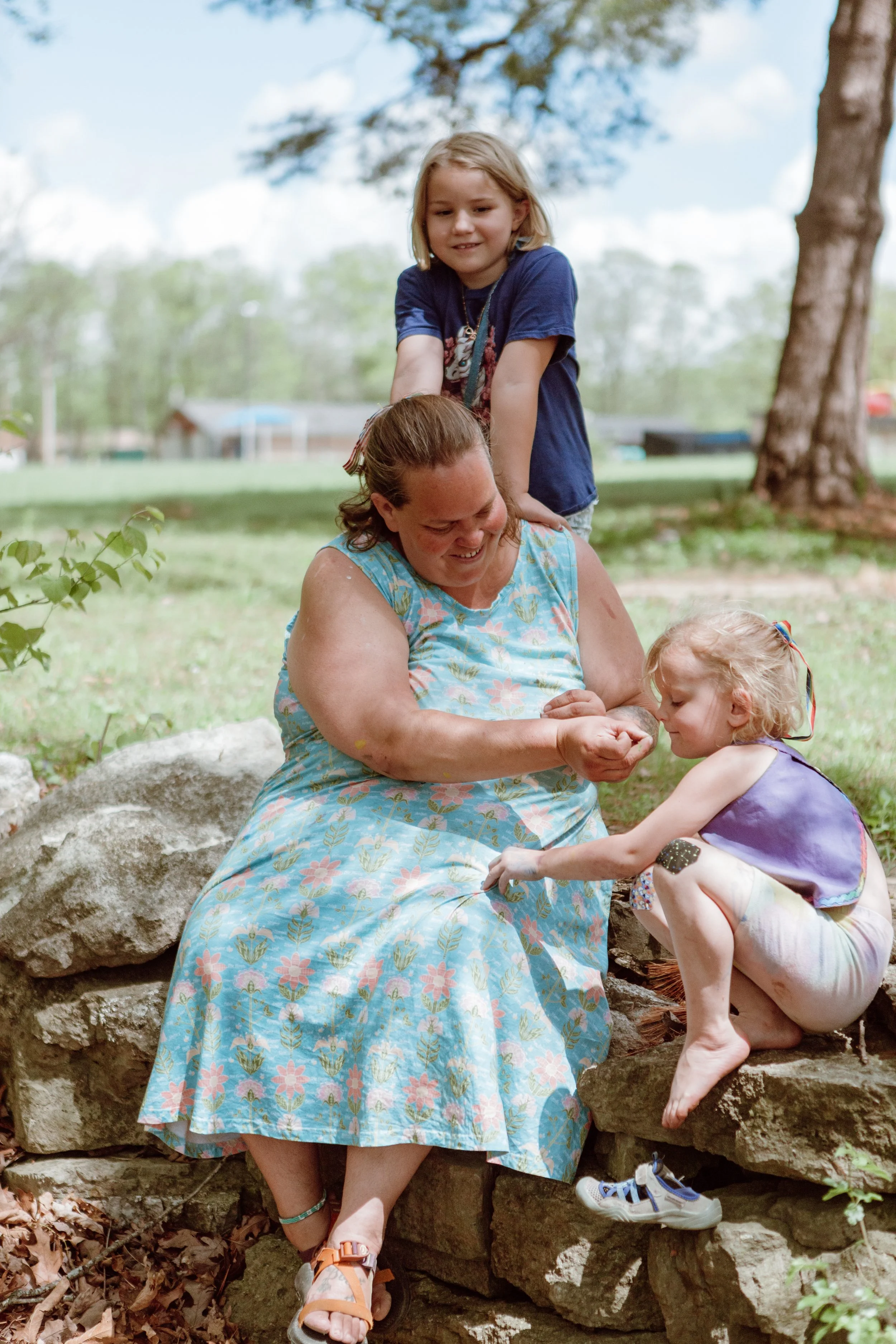 A woman sitting on a rock outdoors, smiling, as three young girls interact with her, one standing behind her and two sitting on the rock, during daytime. The scene takes place in a park or backyard with trees and a cloudy sky.