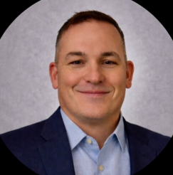 A professional headshot of a man in a suit and light blue shirt, smiling against a gray background.