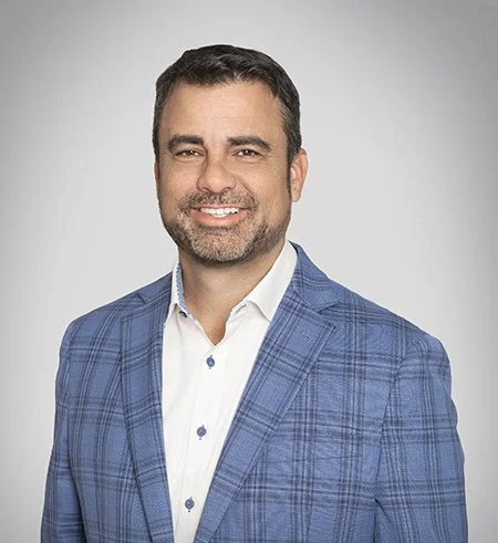 Professional man with dark hair and beard smiling, wearing a blue plaid blazer and a white shirt, against a plain light gray background.