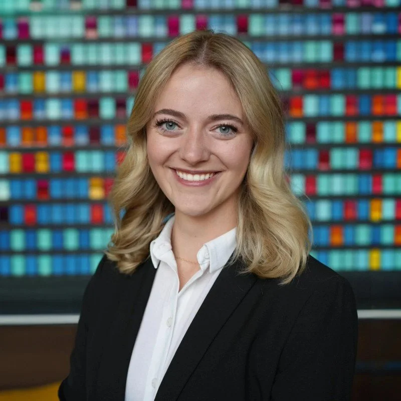 A young woman with blonde hair and blue eyes, smiling, wearing a white shirt and black blazer, standing in front of a colorful background of rows of pills or capsules.