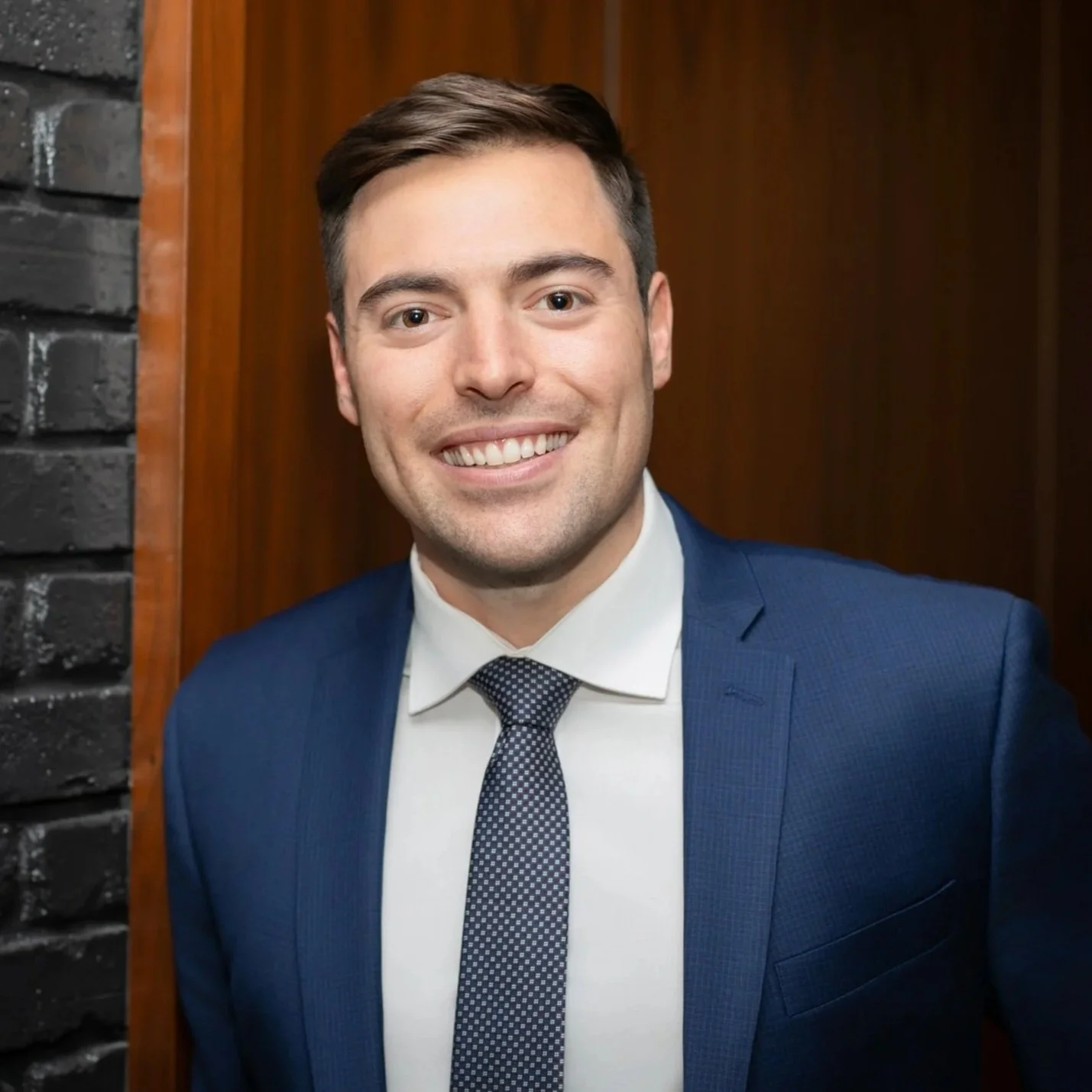 A smiling man in a navy blue suit with a white shirt and patterned tie, posing against a wood-paneled background.