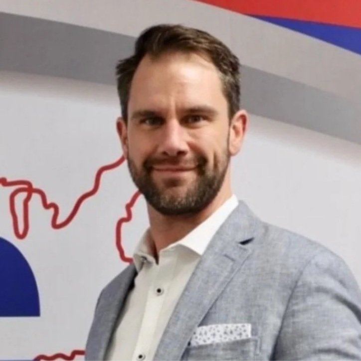 A man with short brown hair and a beard is smiling and wearing a light gray suit jacket with a white shirt. In the background, there is a map outline and a partial logo with blue, red, and gray colors.