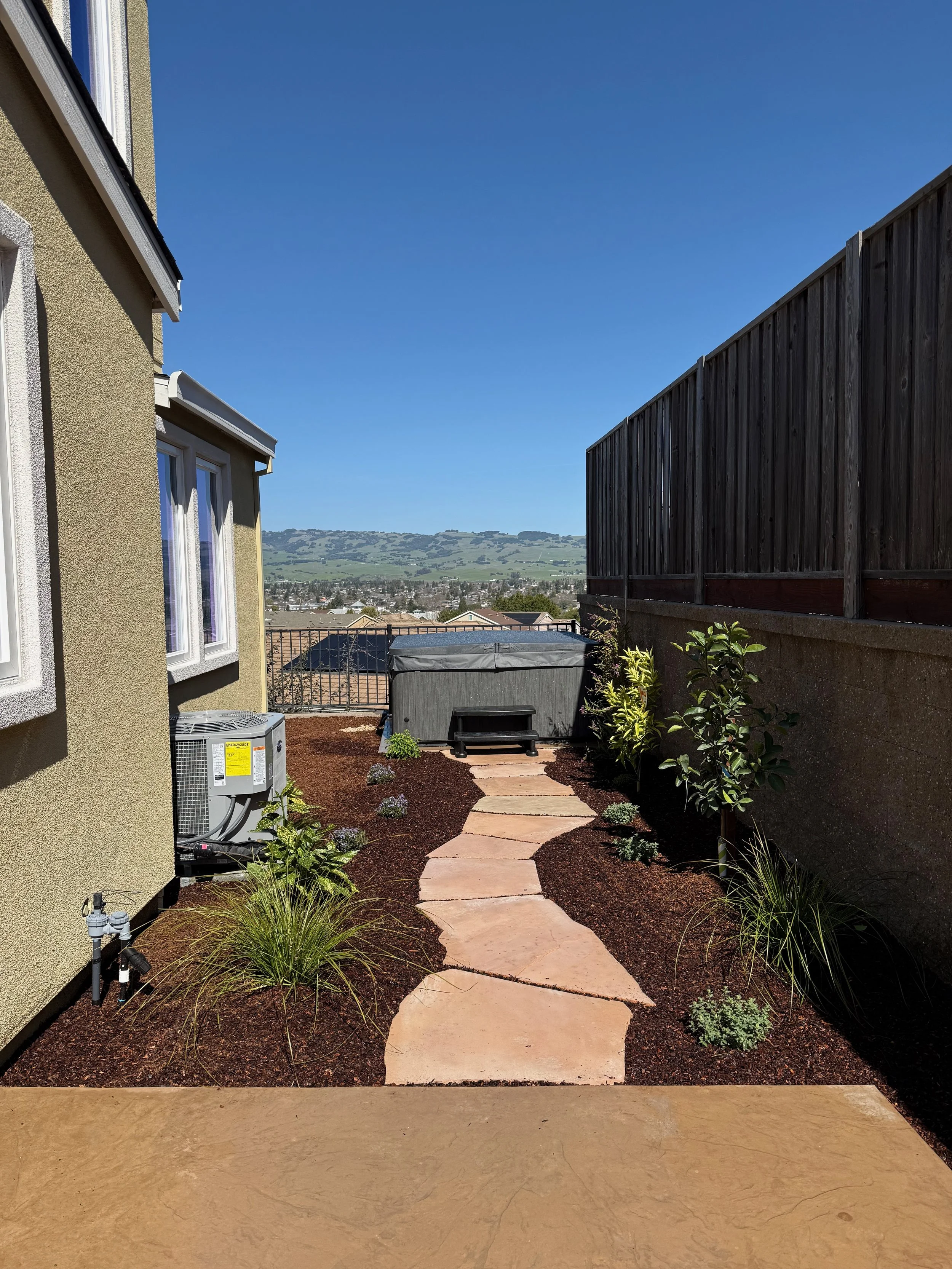 View of a backyard with a stone pathway, plants, a wooden fence, and a view of distant hills under a clear blue sky.