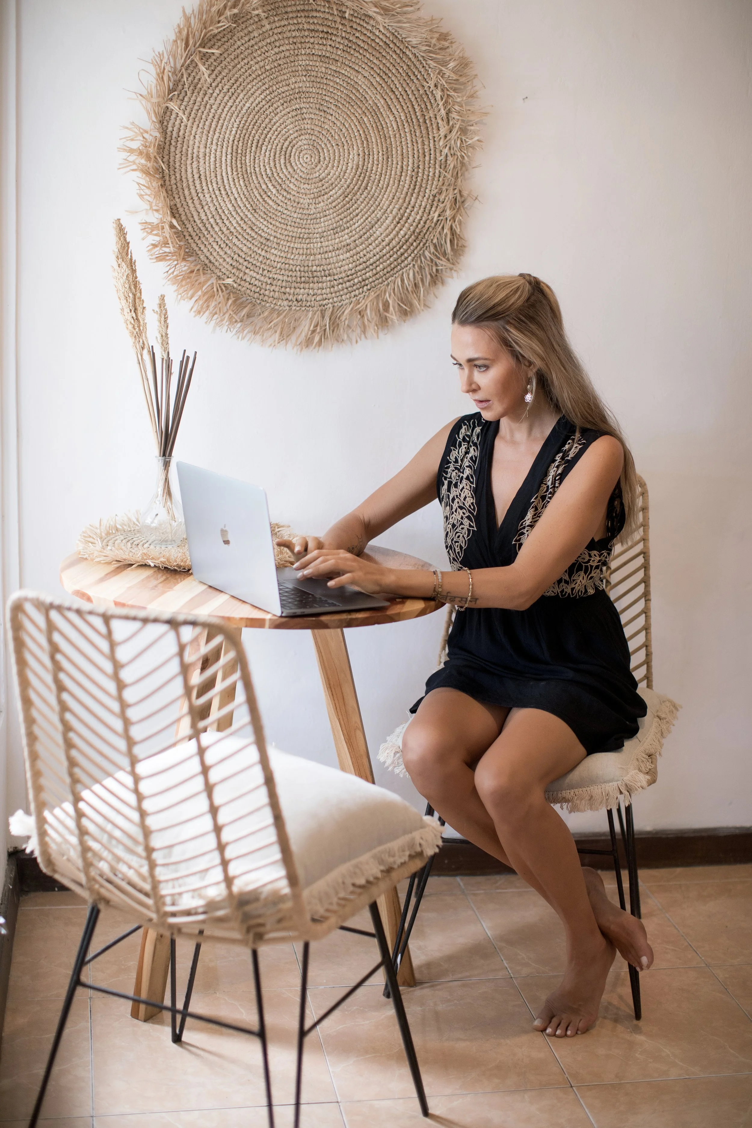 A woman in a black dress sitting barefoot at a small wooden table, working on a MacBook, in a room with a woven wall hanging and decorative dried plants.