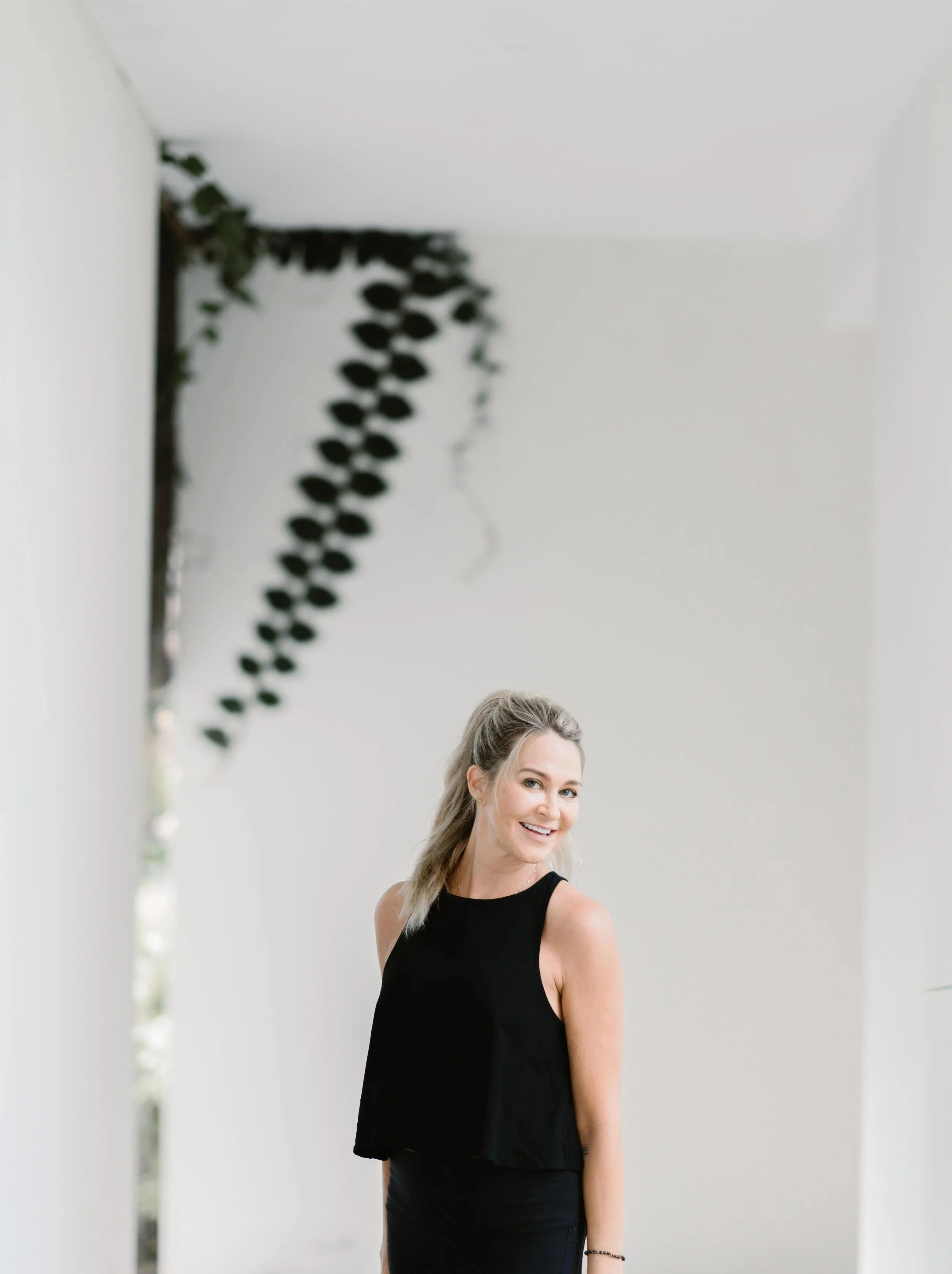 A woman with blonde hair tied back wearing a black sleeveless top, smiling and standing in a white-walled room with hanging greenery.