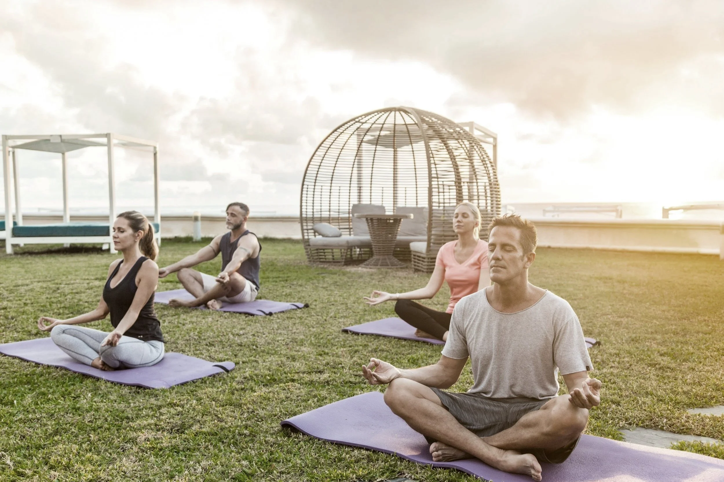 Group of five people practicing yoga on a grassy rooftop at sunset, sitting cross-legged with meditative poses, with outdoor furniture and clouds in the background.