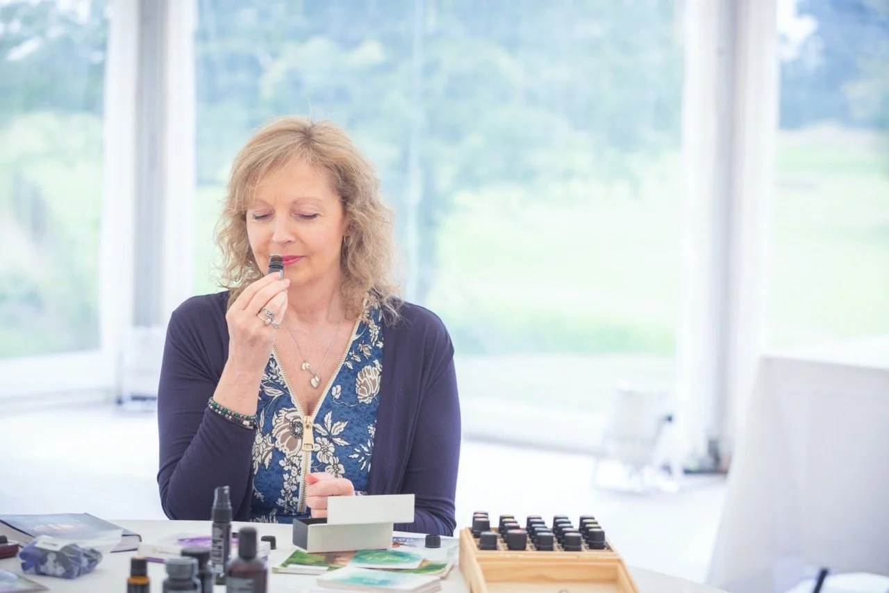 Woman sitting at a table holding a small bottle close to her nose, surrounded by various small bottles and a wooden box with black vials, in a bright room with large windows and greenery outside.