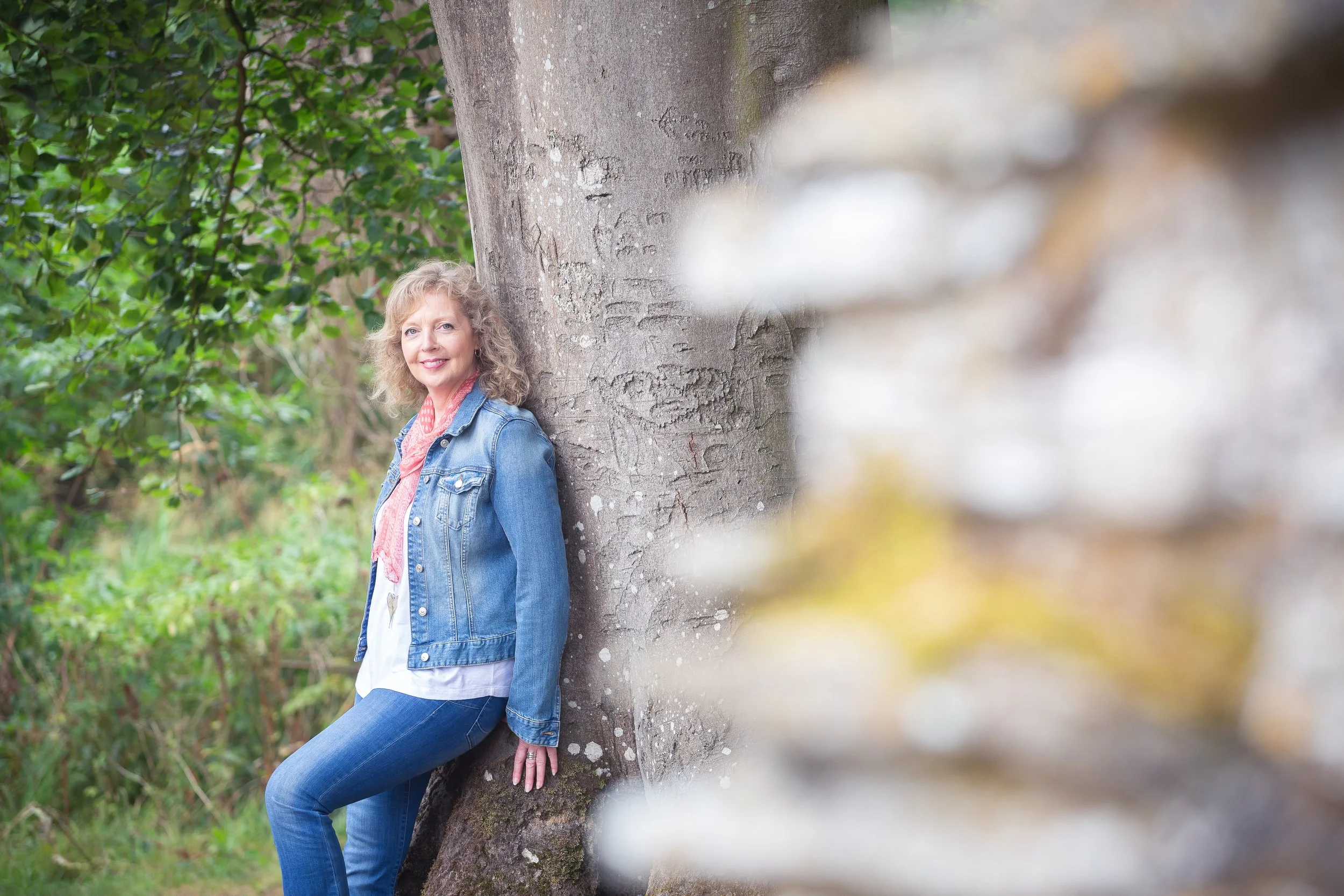A woman with curly blonde hair wearing a denim jacket and jeans standing outdoors next to a large tree trunk, with greenery in the background.