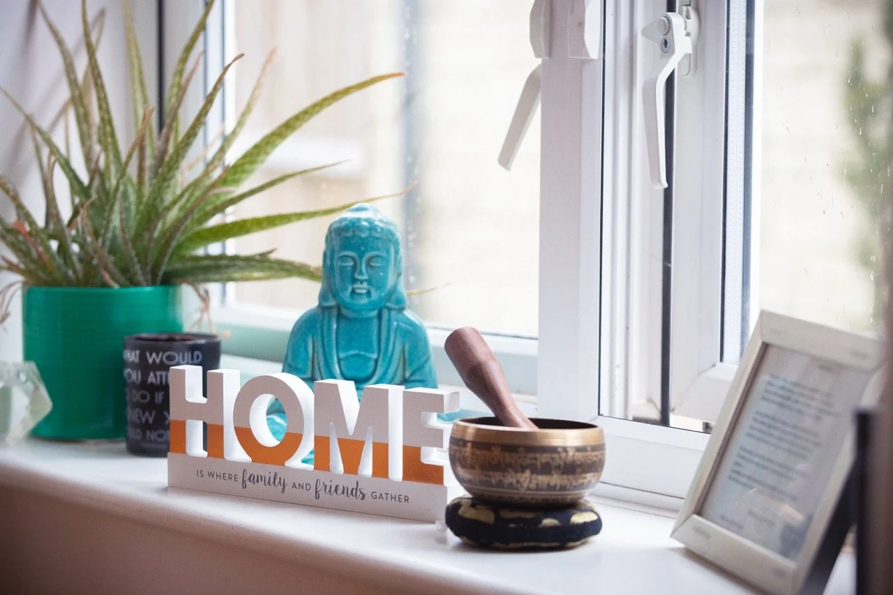 Decorative window sill with a green potted plant, a black mug, a blue Buddha statue, a sign that says 'HOME is where family and friends gather,' a wooden pestle inside a patterned bowl, and a white picture frame.