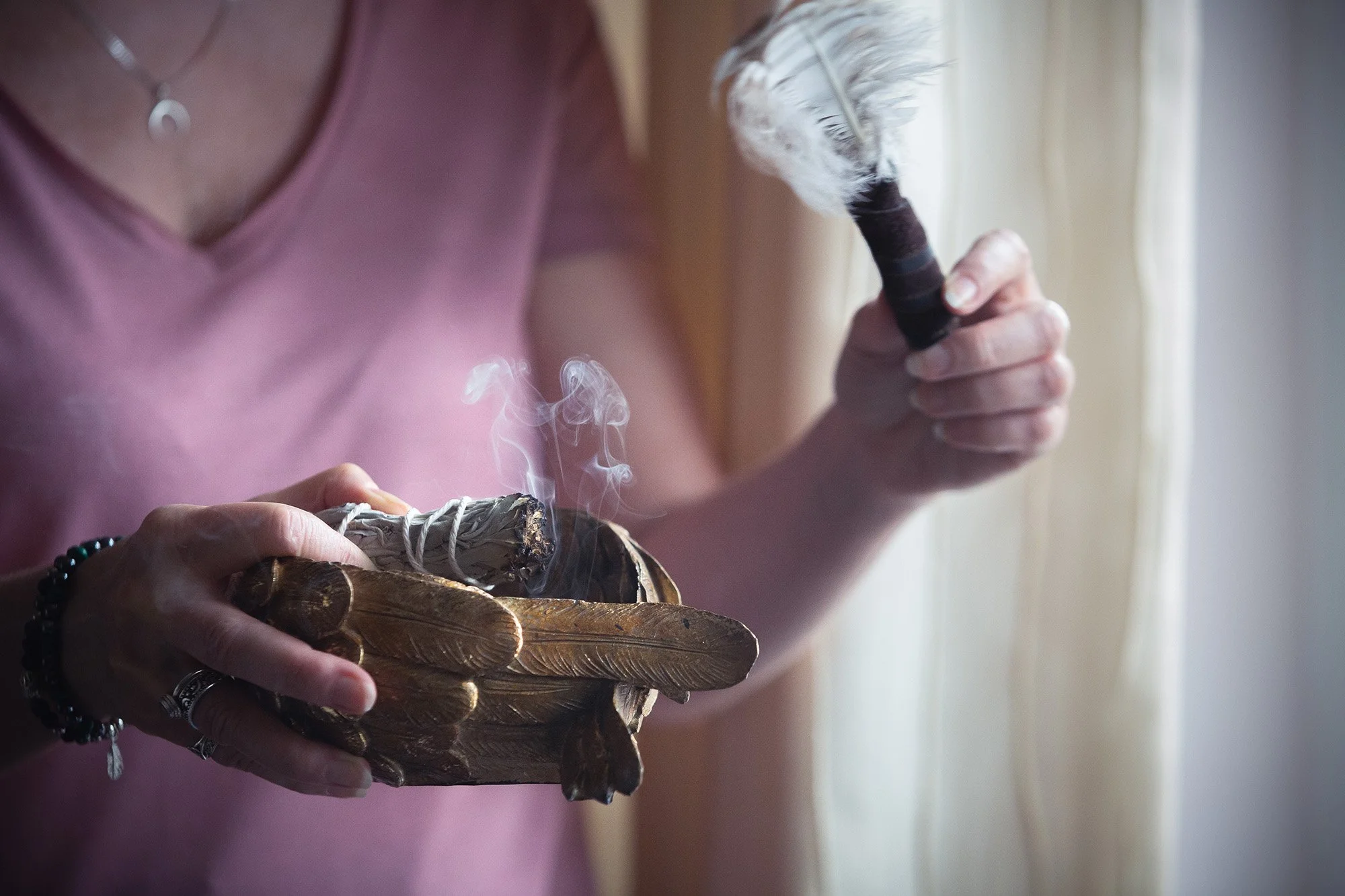 A person holding a burning sage bundle in a wooden bowl and a feather in their hand.