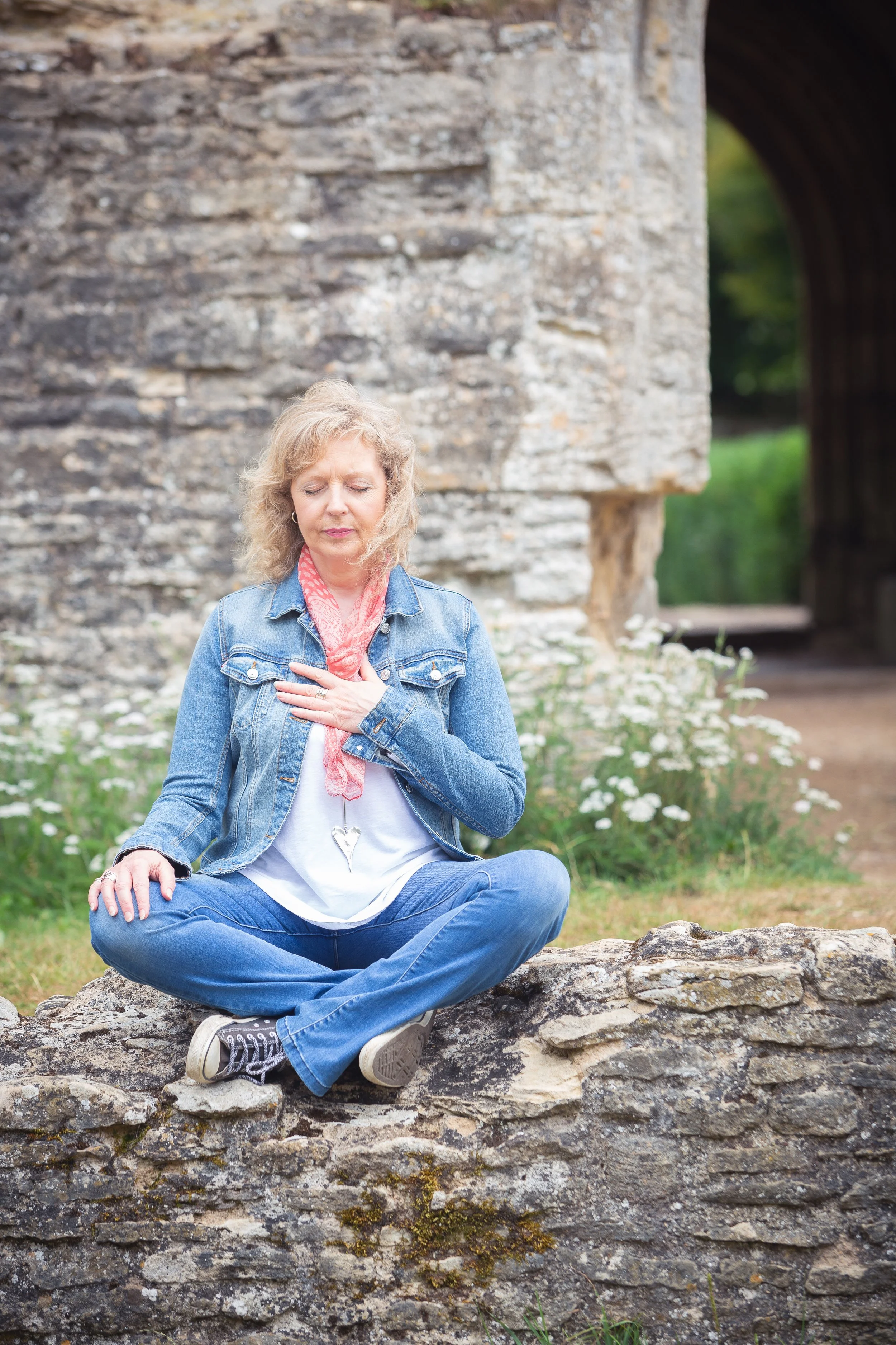 A woman sitting cross-legged on a stone wall, practicing meditation outdoors with her hand over her heart, near an old stone structure and a tunnel in the background.