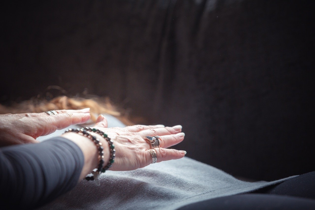 A person's hands resting on their lap, wearing multiple rings and bracelets, against a dark background.