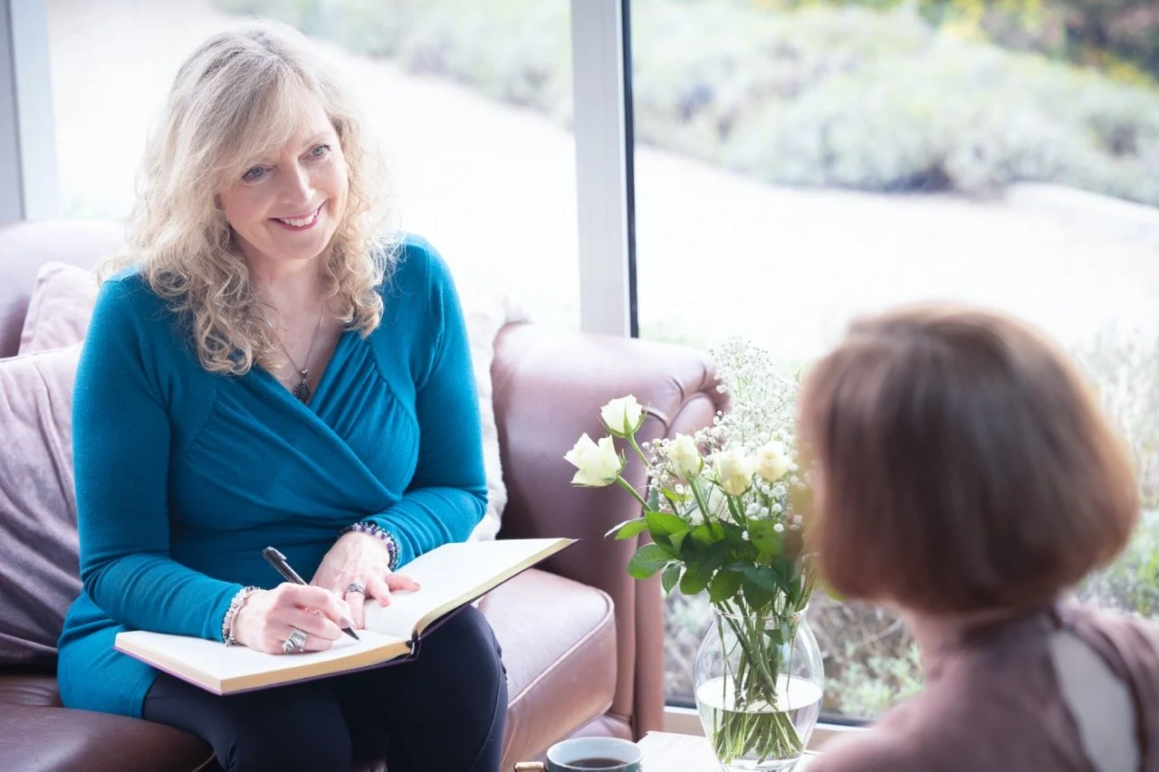 A woman with blonde curly hair, wearing a blue top, seated on a sofa with a notebook, talking to another woman with brown hair. A vase with white flowers is on a table nearby, and a large window with a view of greenery is in the background.