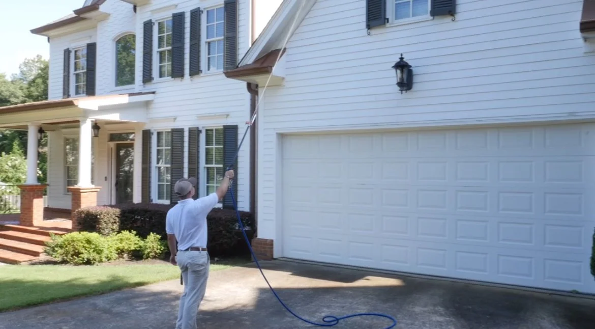 A man in a white shirt and beige pants is cleaning the exterior window of a white house with black shutters using a long cleaning tool, standing on the driveway.