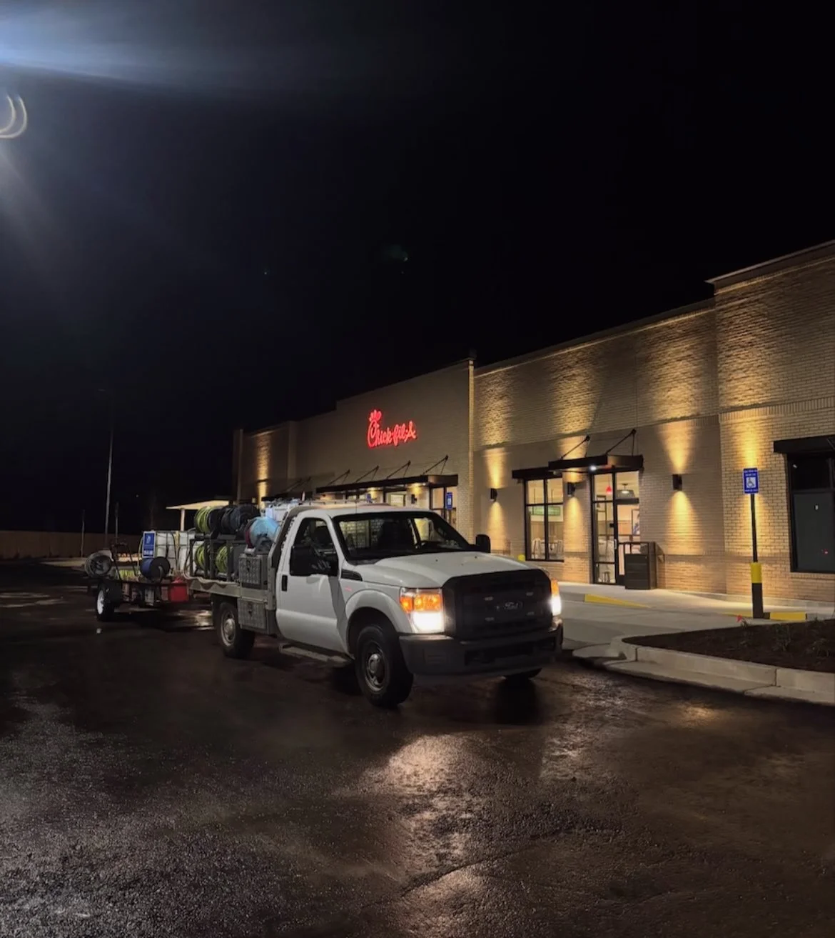 A pickup truck parked outside a Chick-fil-A restaurant at night, with water hoses and equipment loaded in the truck bed.