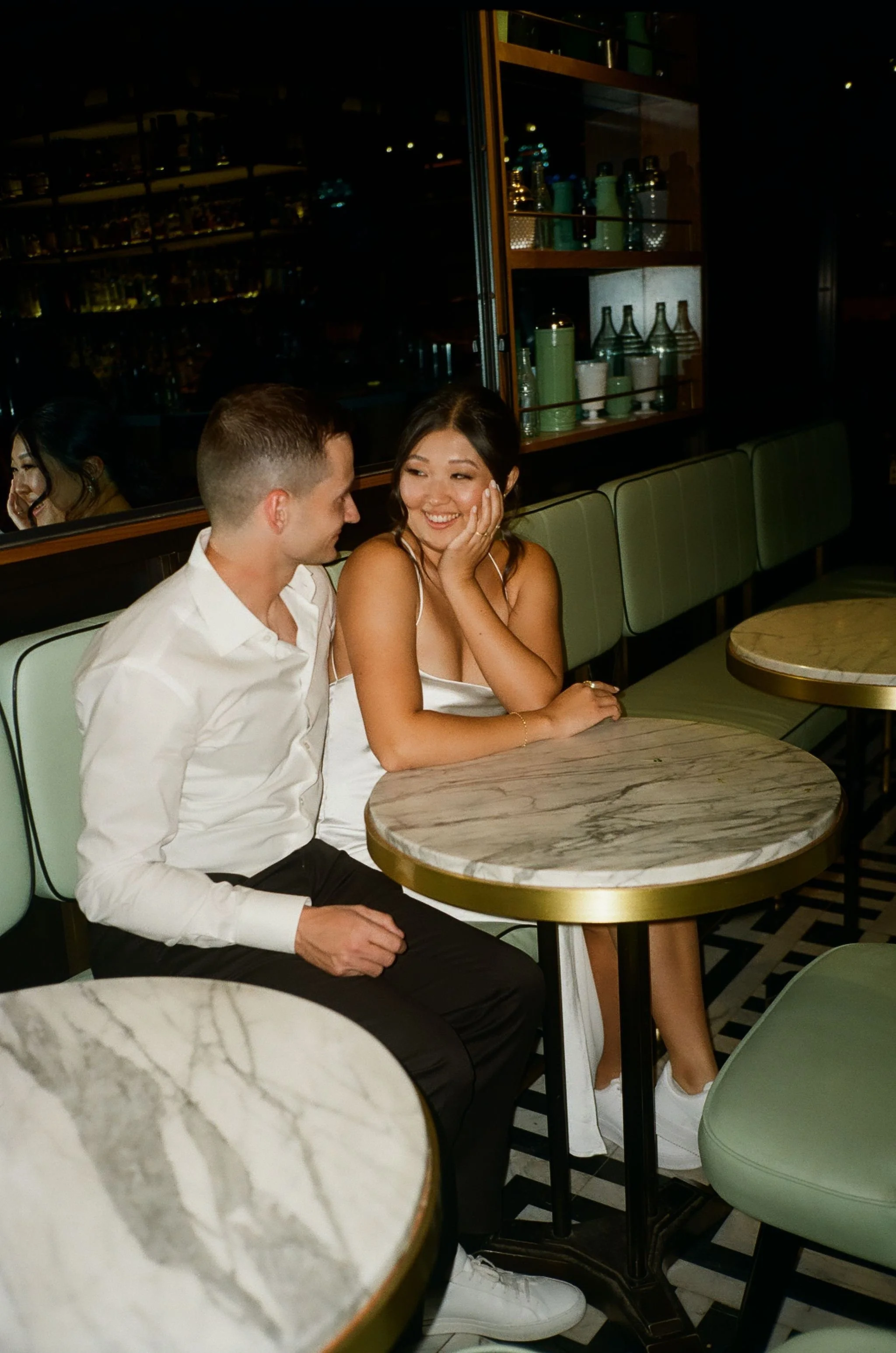 Couple sitting at a marble table in a cafe, smiling at each other romantically, with a reflection in the background mirror.