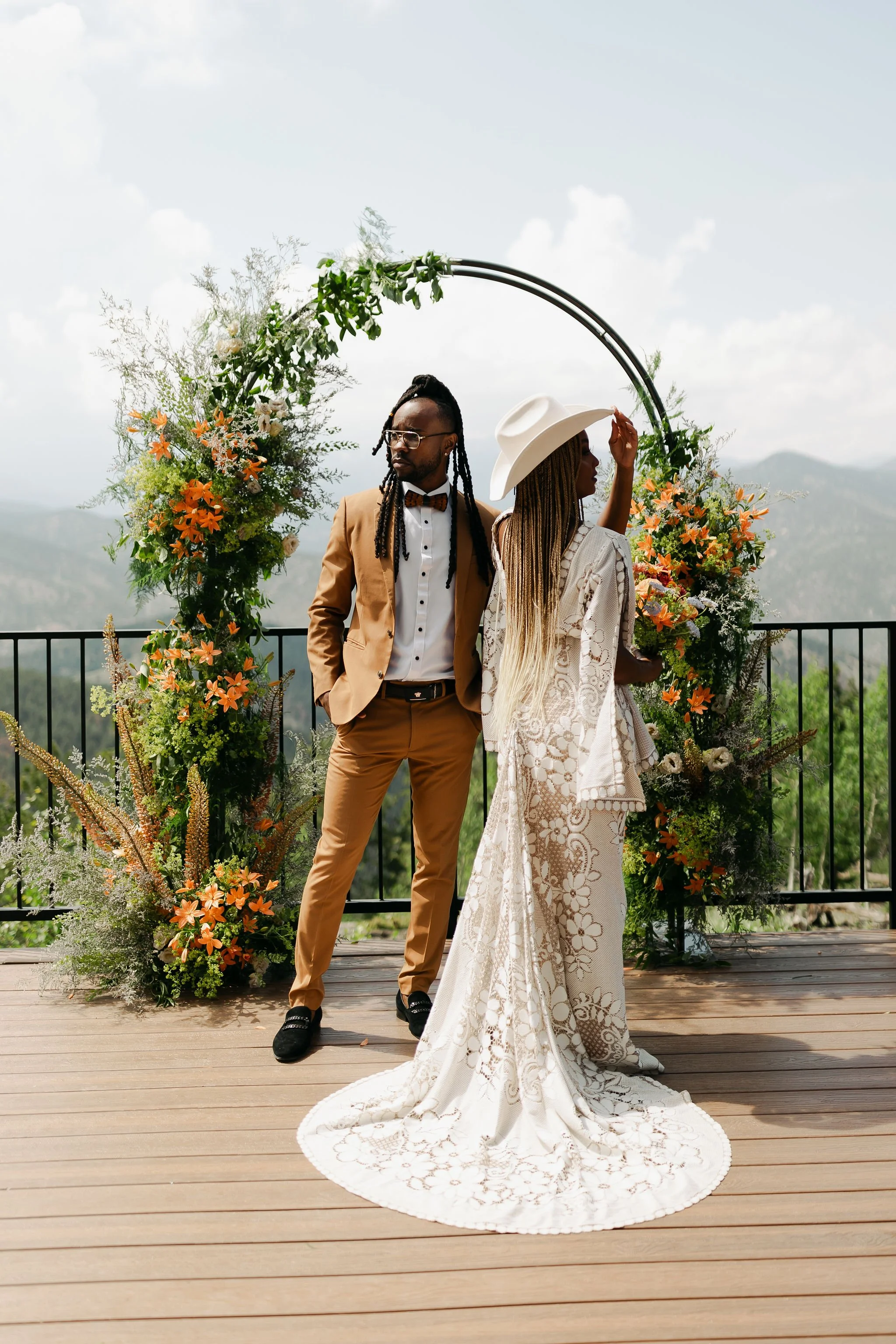A couple standing in front of a floral arch at an outdoor wedding ceremony. The man is dressed in a tan suit with a white shirt and bow tie, and the woman is in a white lace wedding dress with a large white cowboy hat, holding a bouquet.