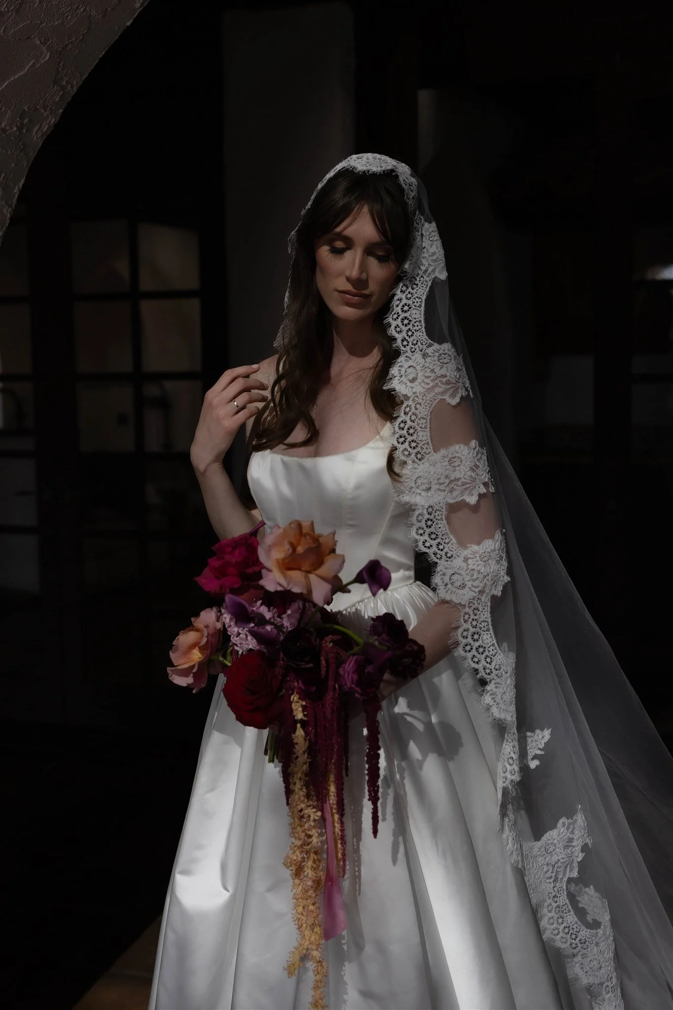 A bride in a white gown holding a bouquet of various colored flowers, with lace details on her veil, standing in a dimly lit room.