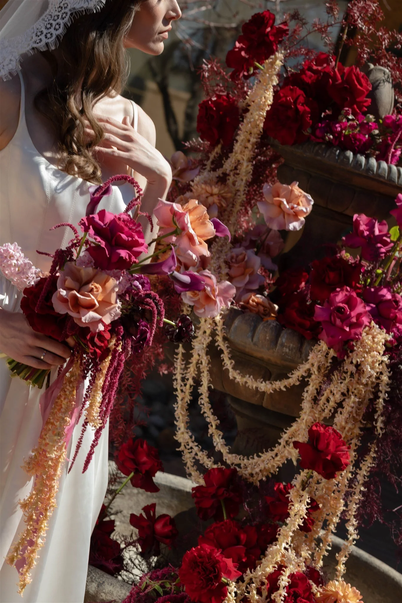 A bride in a wedding dress and veil stands near a fountain decorated with pink, peach, and red flowers, including cascading floral garlands, at an outdoor wedding ceremony