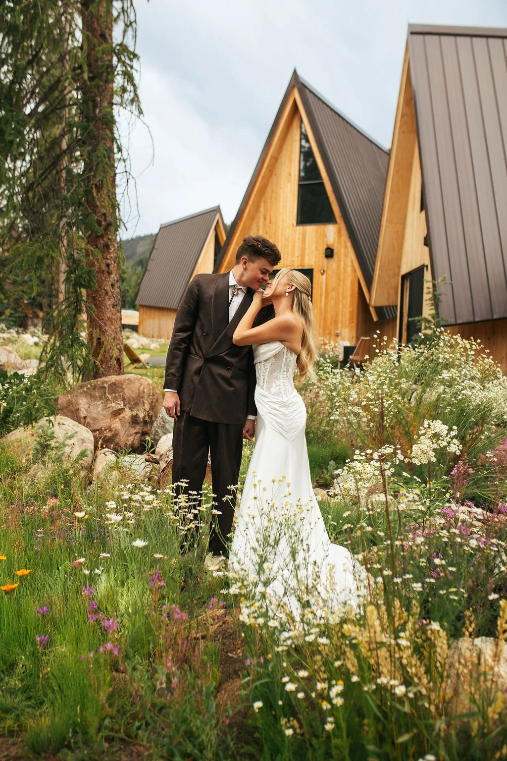 A couple in wedding attire standing close together outdoors, surrounded by wildflowers and rocks, with modern wooden houses in the background.
