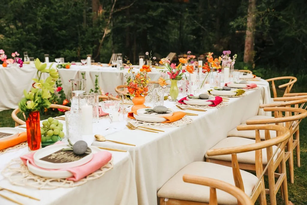 Long outdoor dining table set for a celebration with colorful flowers, pink and orange accents, and natural wood chairs in a forest setting.