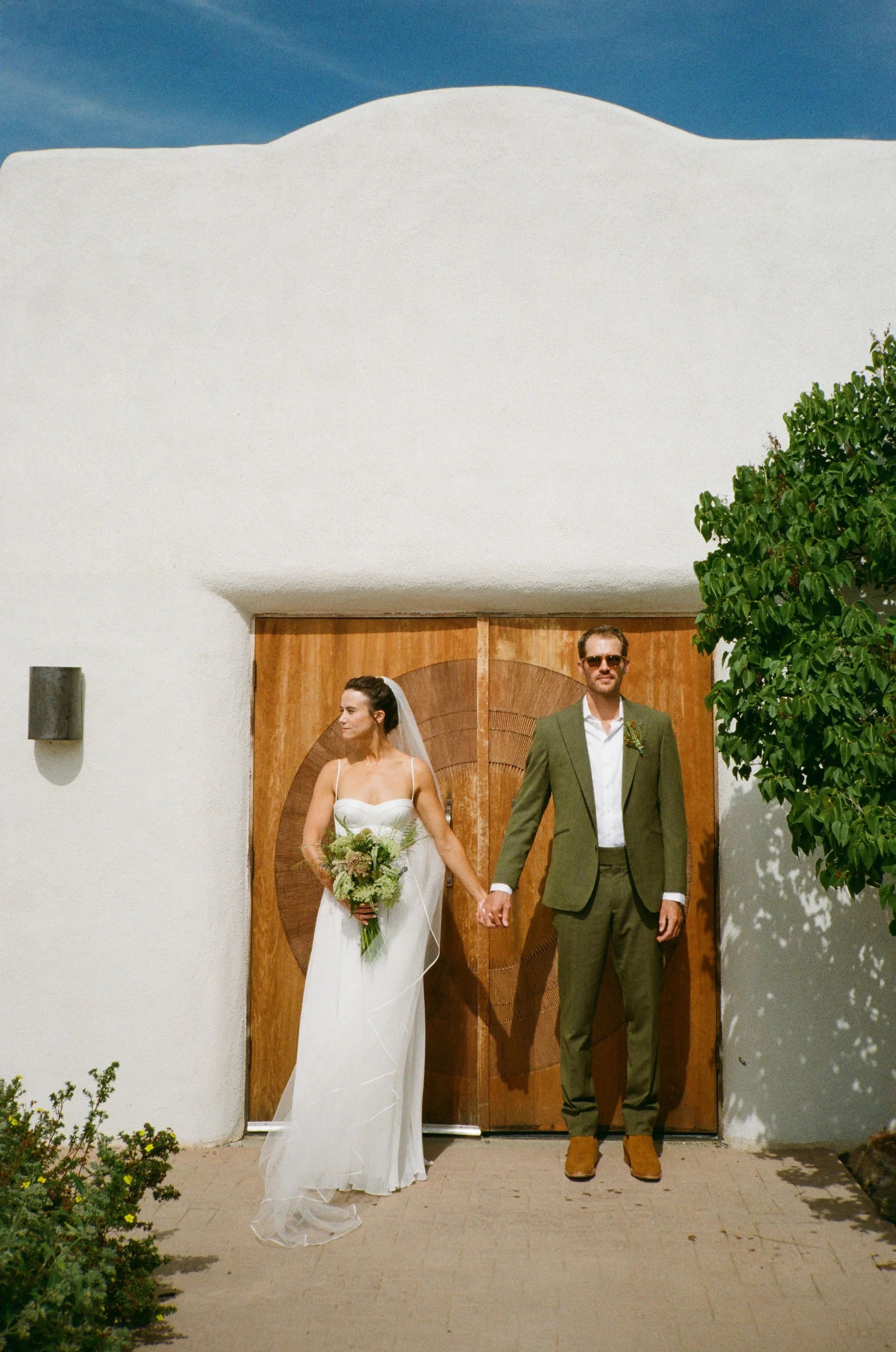 A bride and groom holding hands in front of a wooden door during a wedding, with white stucco walls and clear blue sky.
