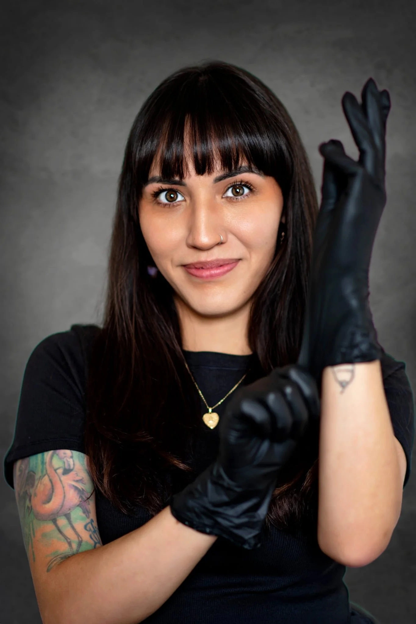 A woman with long dark hair, brown eyes, and a nose ring, wearing a black glove on her right hand, a black shirt, gold necklace with a heart pendant, and visible tattoos on her upper left arm, smiles at the camera against a gray background.