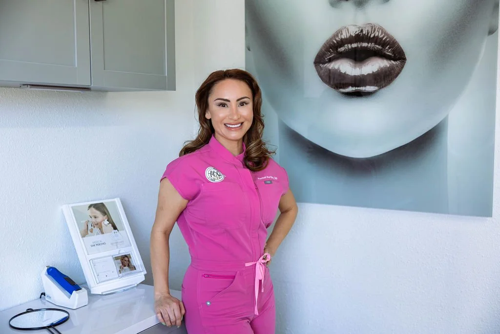 A smiling woman wearing pink medical scrubs standing in front of a wall with large black-and-white art of lips and nose. She has brown hair and is posing with one hand on her hip.