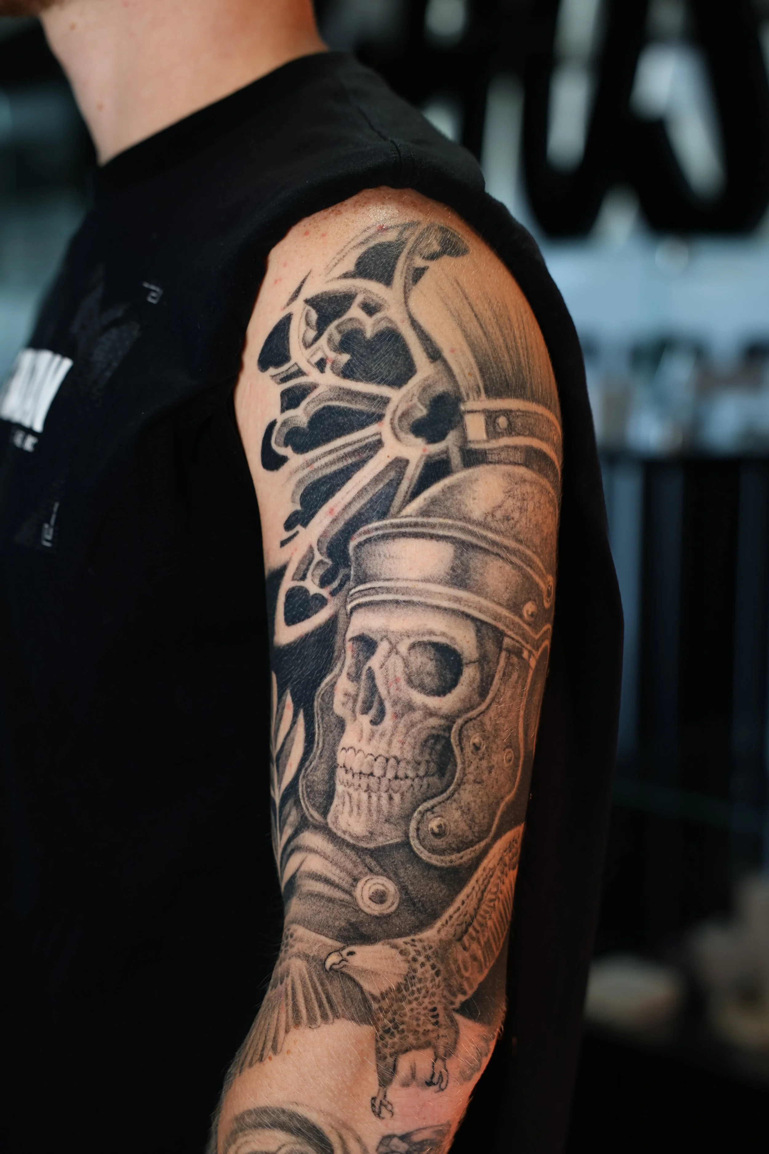 Close-up of a person's upper arm and shoulder with a detailed black and gray tattoo of a skull with mechanical or steampunk elements, including gears and cogs, extending from the shoulder down to the forearm.