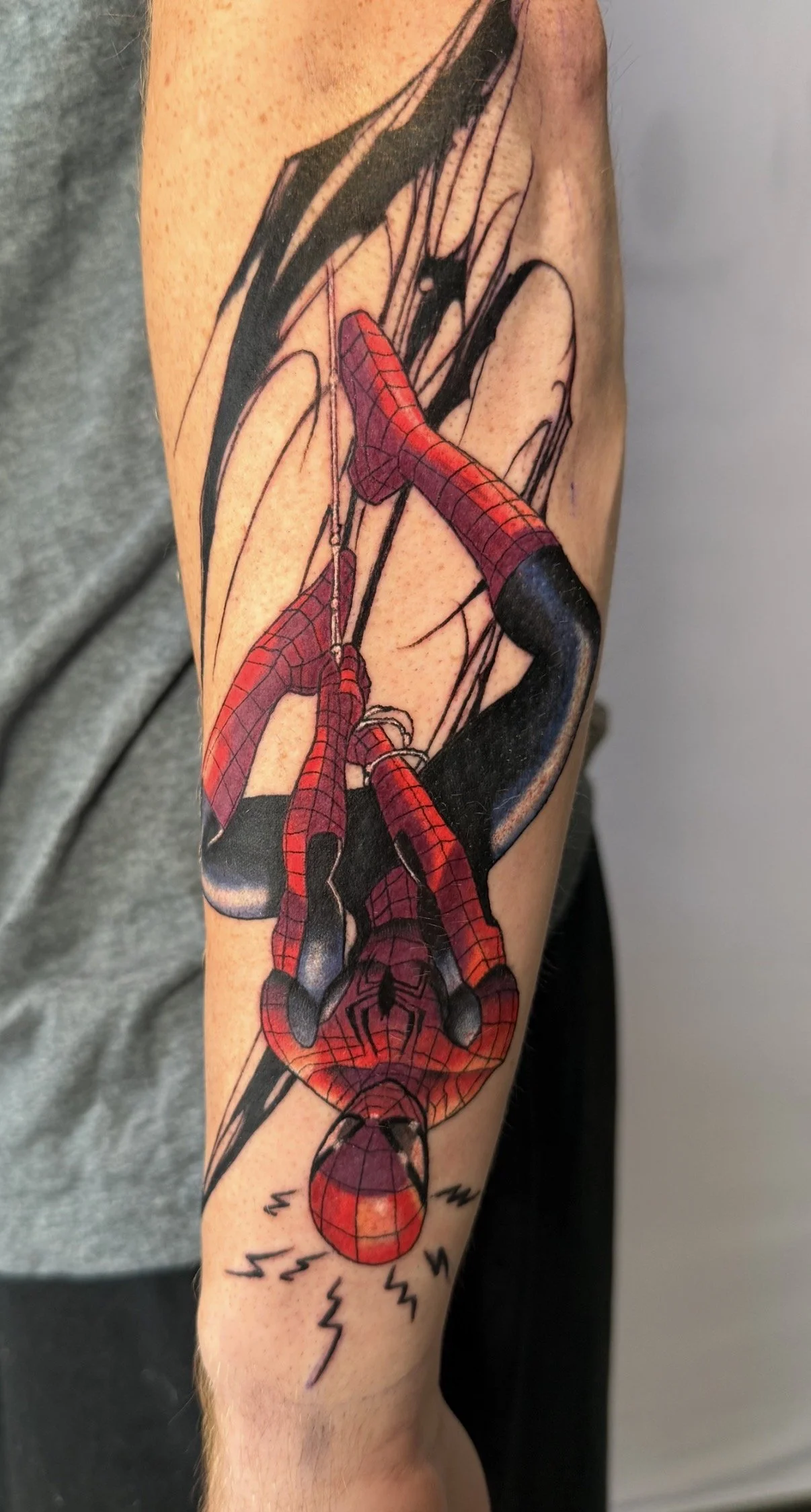 Colorful tattoo of Spider-Man hanging upside down, with comic-style background and lightning bolt details.