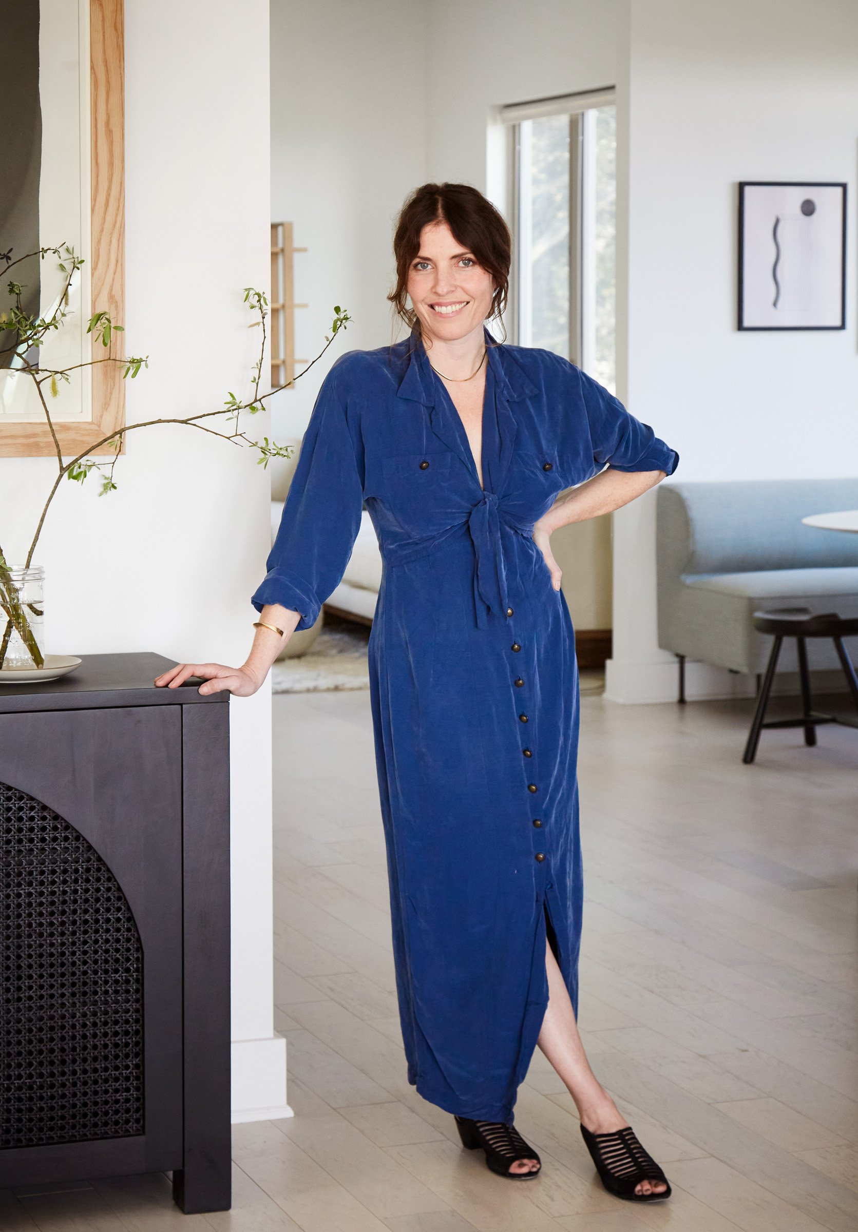 A woman in a blue dress standing in a modern living room, smiling, with her hand on a dark cabinet.