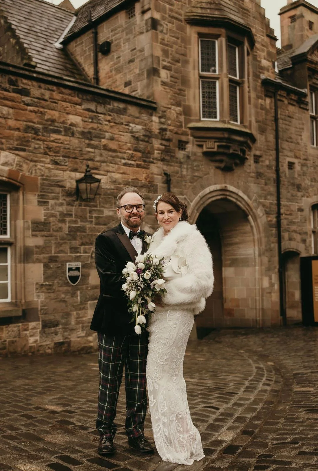 A bride and groom standing together outside a historic stone building on their wedding day, smiling at the camera. The groom is wearing a tuxedo with plaid pants, and the bride is dressed in a lace wedding gown with a white fur coat, holding a bouque