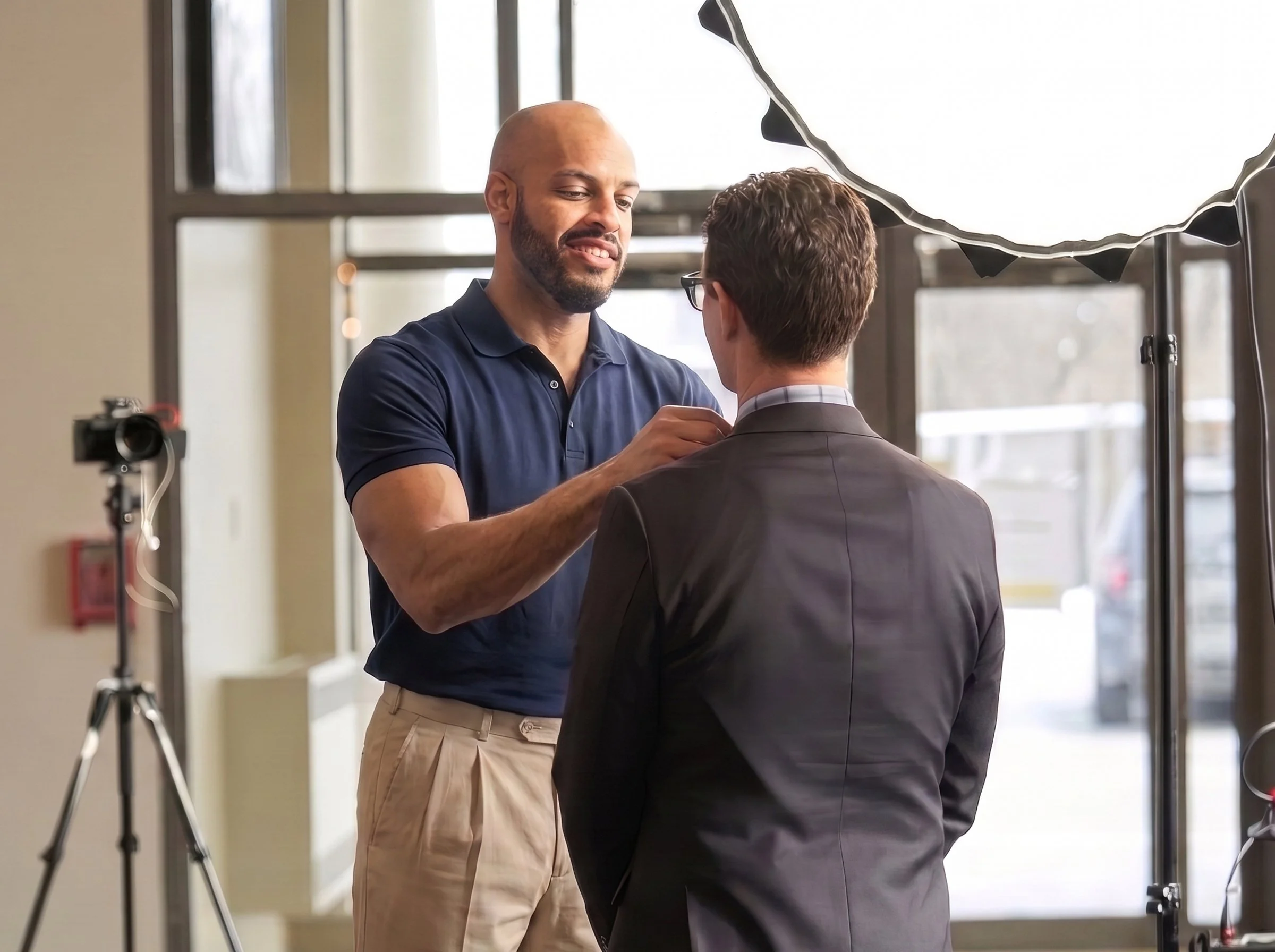 A man with a beard and short hair, wearing a blue polo shirt, helps a man with glasses and brown hair in a suit adjust the collar of his shirt in a well-lit indoor space with large windows. There is photography equipment, including a camera on a tripod and a large softbox light, indicating a photo shoot setting.
