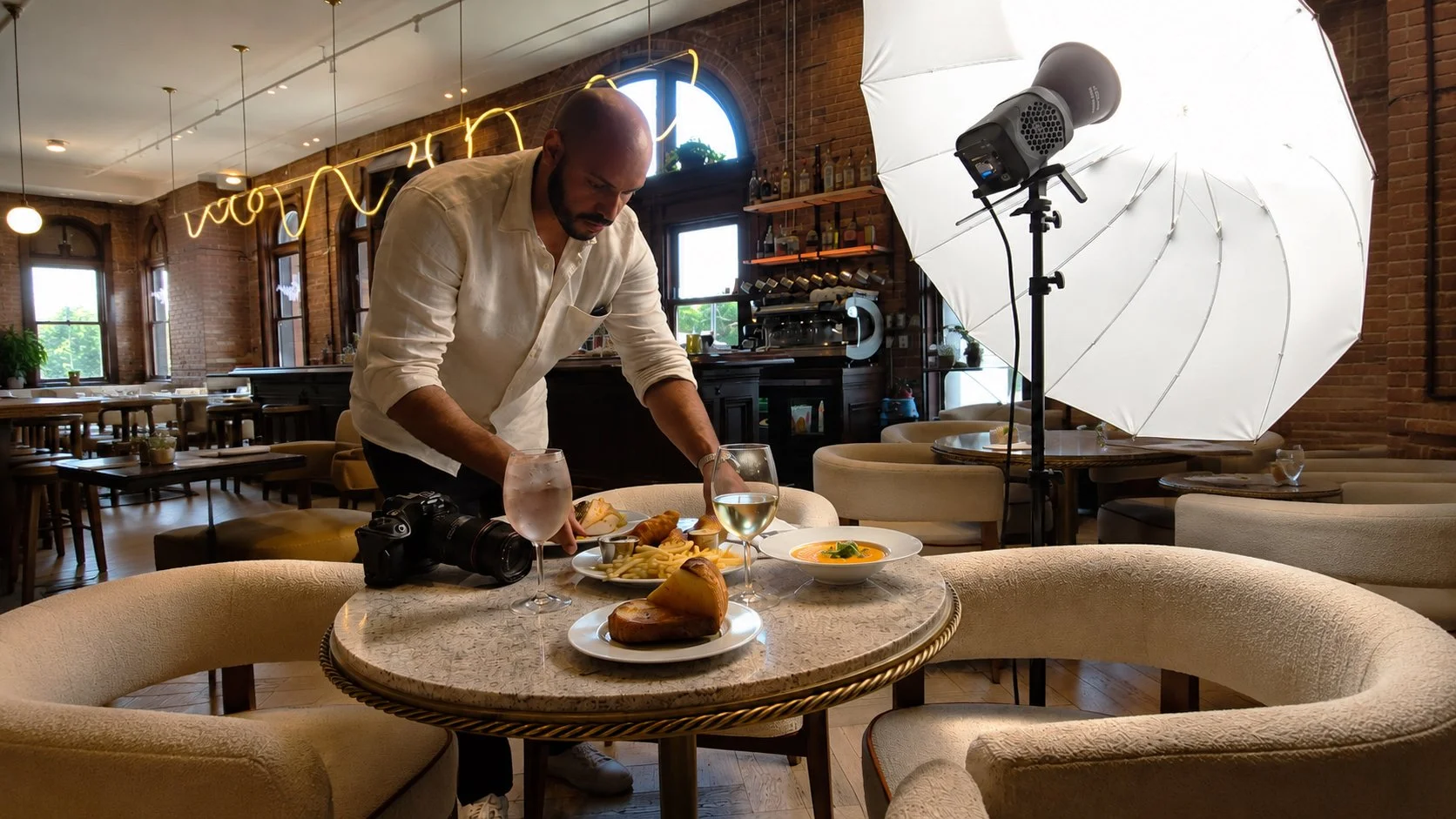 Photographer setting up food on a table in a restaurant with large windows, brick walls, and a white umbrella-style lighting setup.