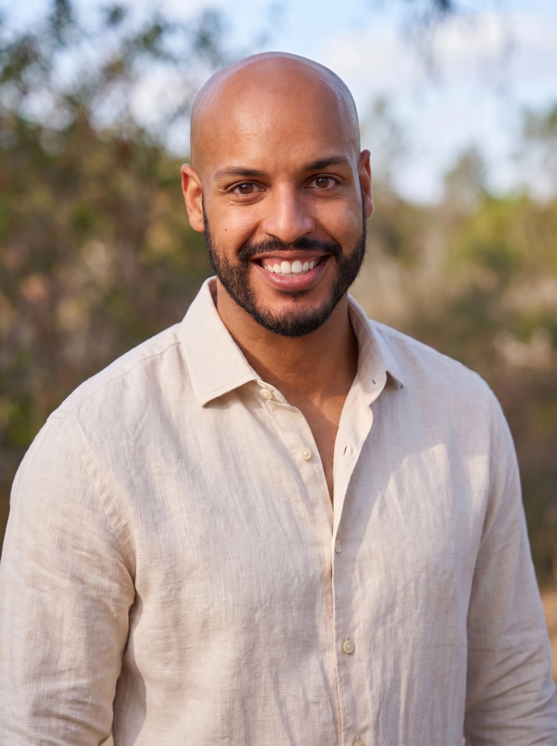 A smiling man with a bald head and beard wearing a light-colored button-up shirt, standing outdoors with blurred trees and sky in the background.