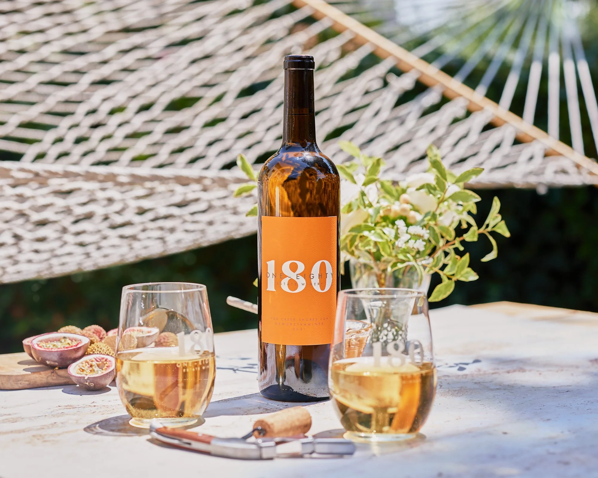 A bottle of wine with a bright orange label marked with the number 100, surrounded by glasses of white wine, passion fruit halves, and a small bowl of fruit on a wooden table outdoors, with a background of greenery and a canopy.