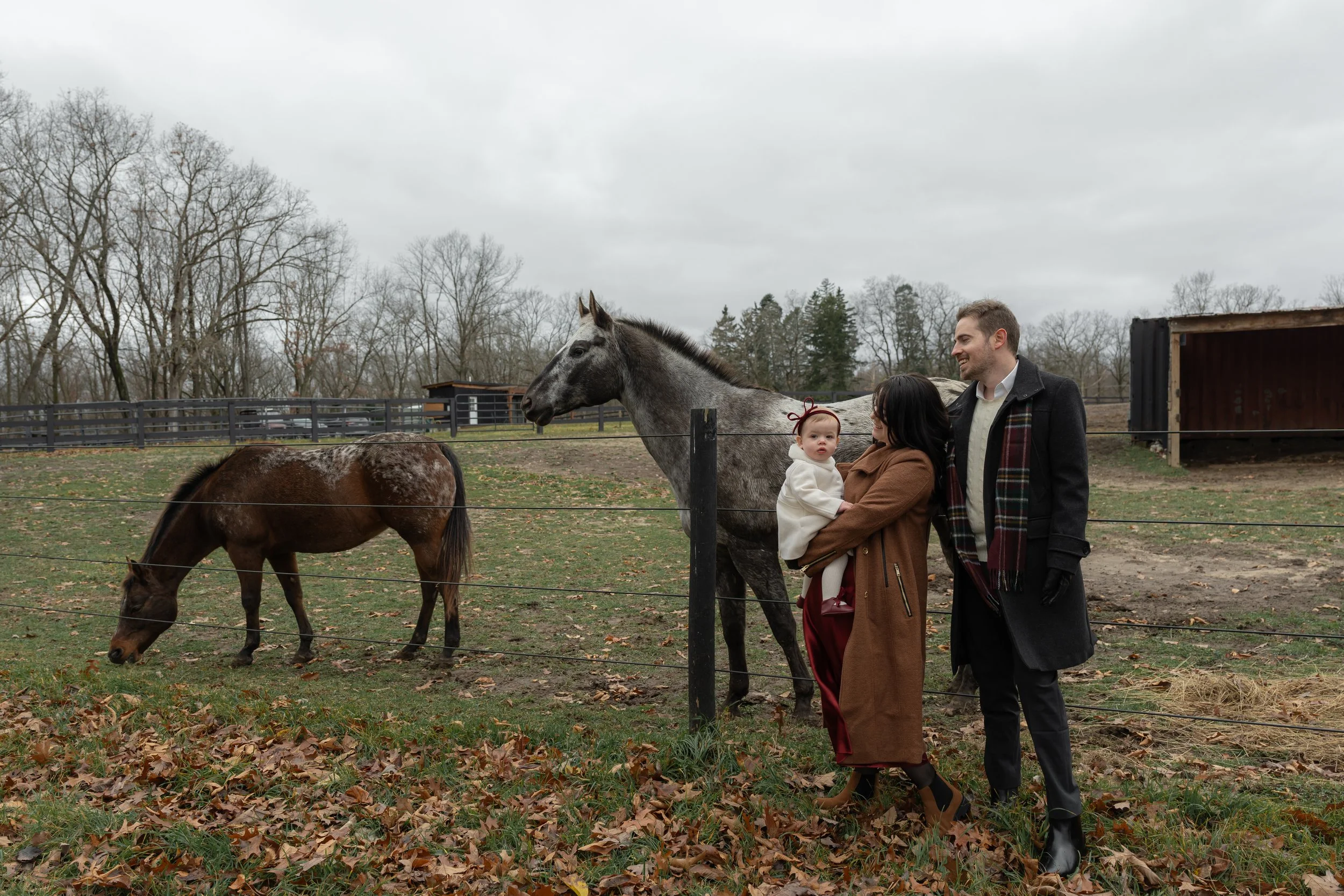 A family of three at a horse farm on a cloudy day, with two horses in the background. The woman is holding a baby girl, and a man stands beside them, all smiling and dressed warmly.