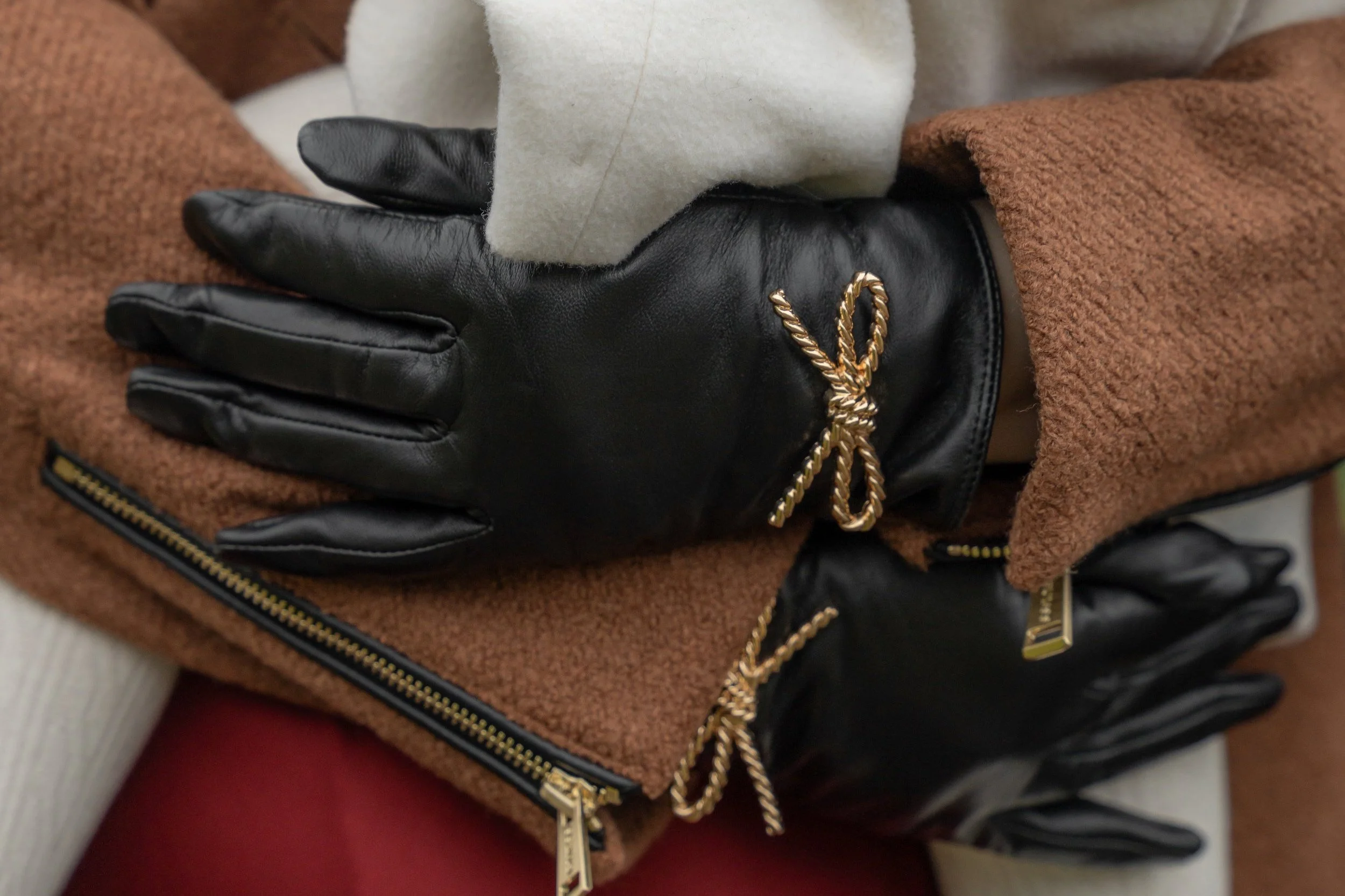 Close-up of a person's hand wearing a black leather glove with a gold-colored chain detail, resting on a brown fleece jacket with a black zipper.