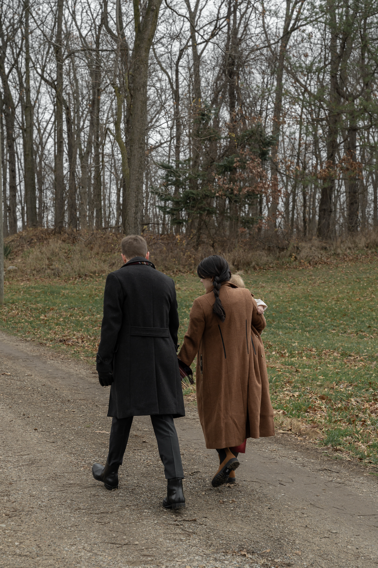 A man and woman walking together on a dirt path through a park or forest, with trees and fallen leaves in the background on a cloudy day.