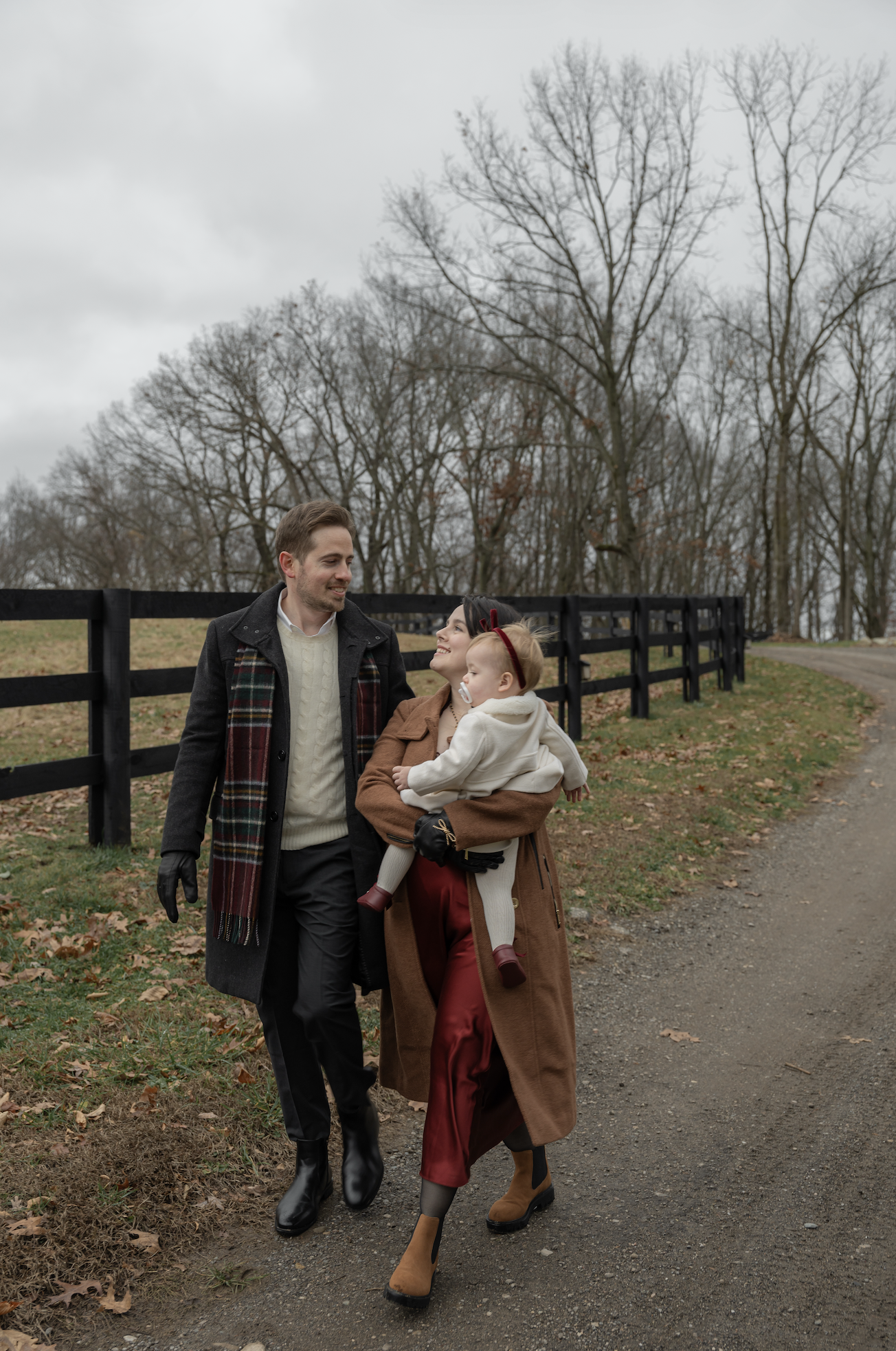 A family of three walking outdoors on a cloudy day, with leafless trees and a wooden fence in the background. The man is wearing a long coat with gloves, the woman is holding a young girl, and the girl has a pacifier and a headband. All are dressed w